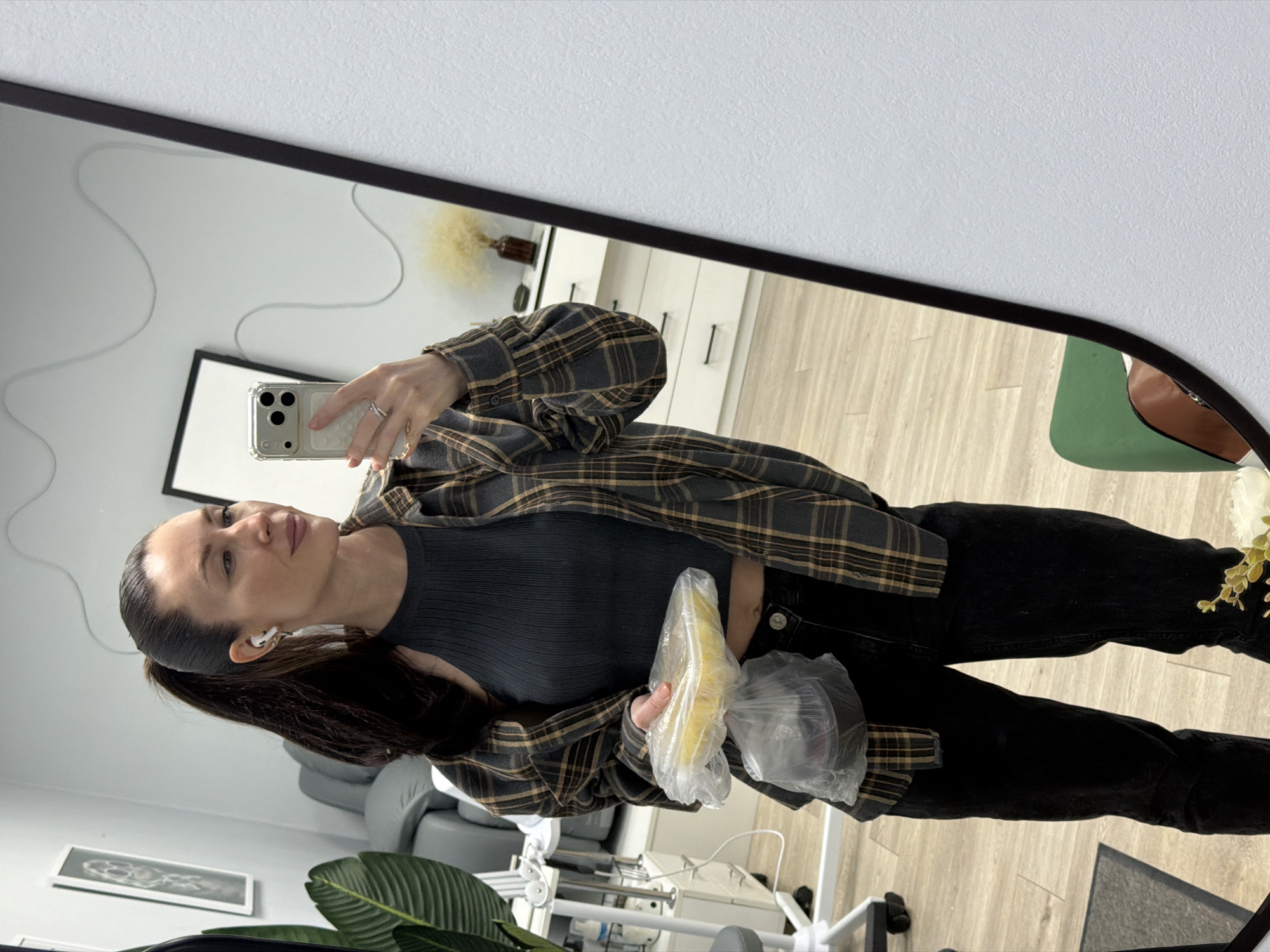 A woman is taking a selfie in a mirror. She is wearing a gray ribbed top, black jeans, and a gray and brown plaid flannel shirt. She is holding two plastic bags, one containing what appears to be food. She has her hair pulled back in a ponytail and is wearing an earpiece. The reflection in the mirror shows a room with medical equipment and furniture. Based on surrounding metadata, this image was taken in the city of Perm, Russia.