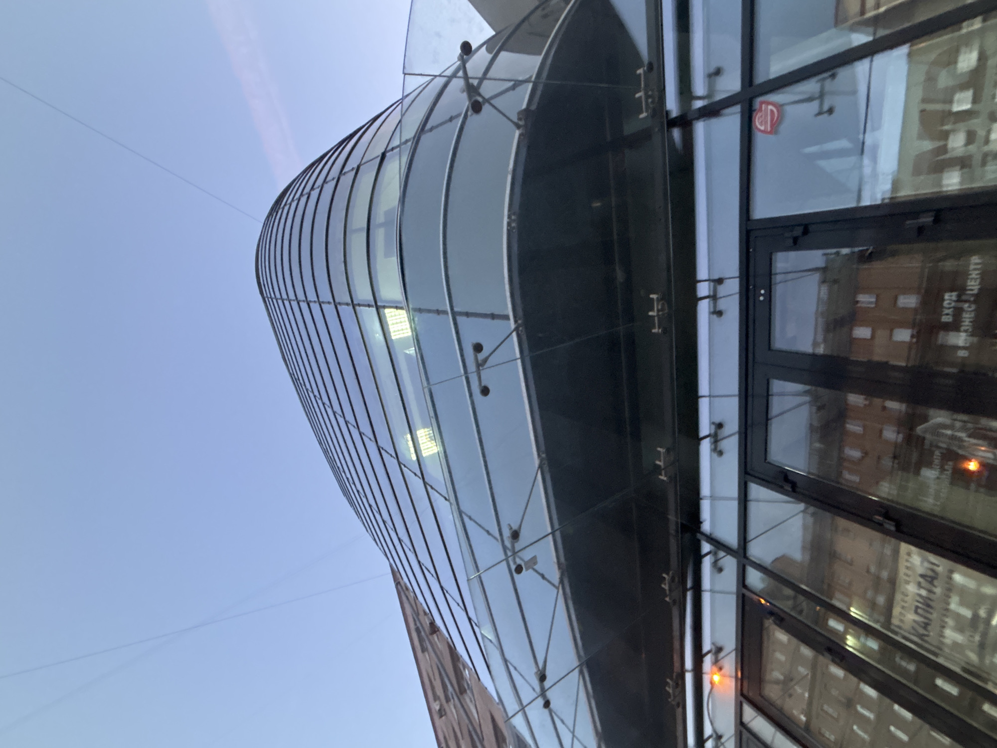 This is a low-angle, full-shot photograph of a modern glass-fronted building in Novosibirsk, Russia. The building has a curved facade with horizontal window panes that reflect the sky, which is a pale blue with faint wisps of clouds. A glass railing with metal supports is visible on an upper level. Below this, the entrance to the building features large glass doors and windows that reflect the surrounding architecture. Text is visible through the glass, including "КАПИТАЛ" which translates to "Capital," and "ВХОД В БИЗНЕС ЦЕНТР" meaning "Entrance to business center." There are no people visible in the frame. The overall scene suggests an urban environment, likely during daylight hours given the brightness of the sky and reflections.