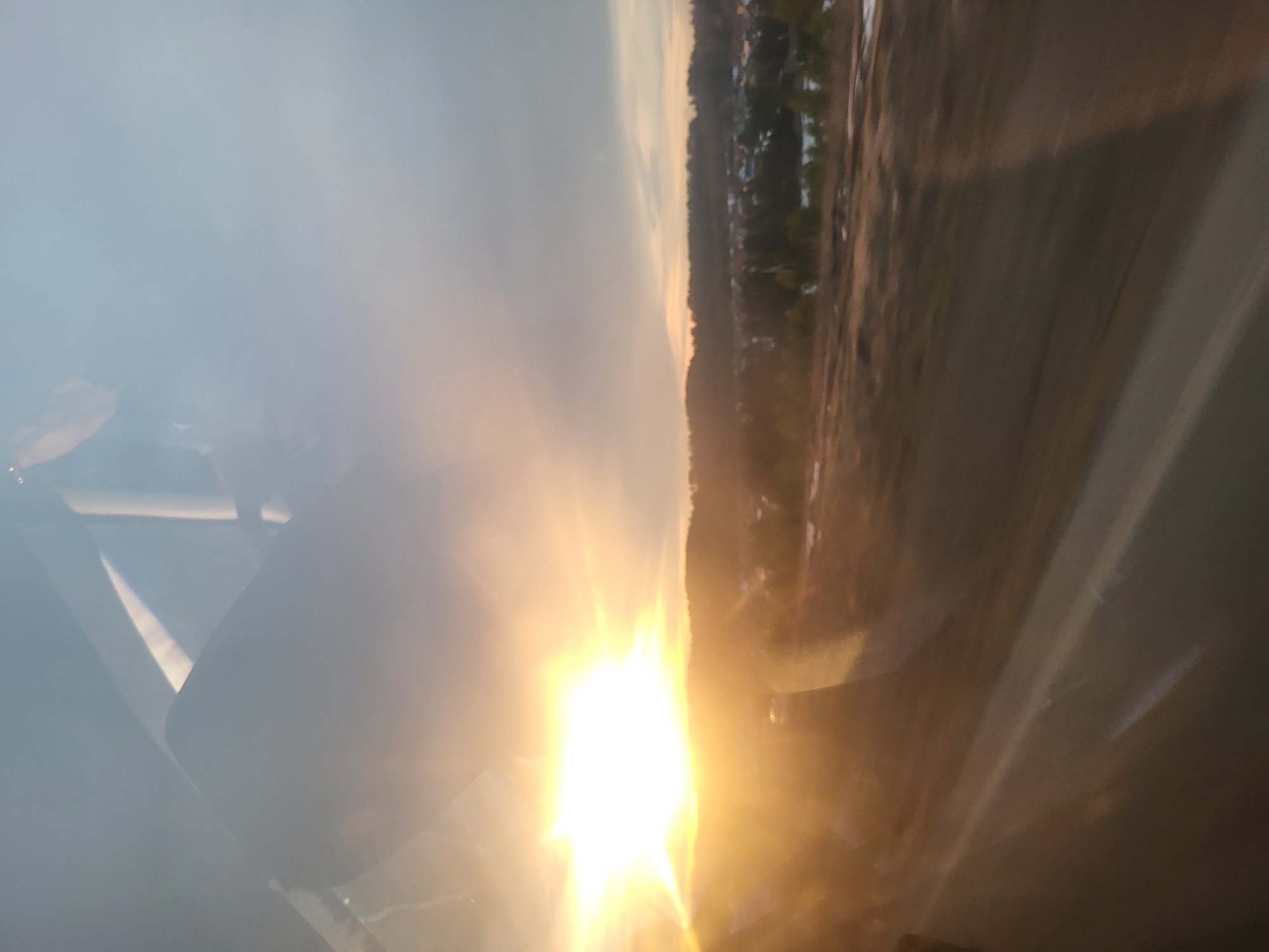 The image appears to be taken from inside a vehicle, possibly an airplane, given the presence of what seems to be a wing strut and the angle of view. The sun is low on the horizon, causing glare and obscuring details in the foreground. The landscape visible outside the vehicle features a mix of vegetation and what could be buildings in the distance. The specific location within Фоки, Russia, cannot be determined from this image alone due to the limited detail and glare.