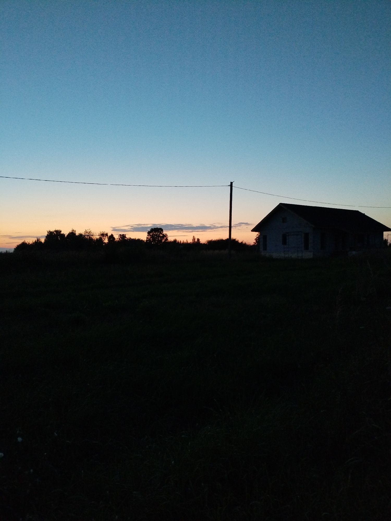 An outdoor, eye-level, full-shot photograph captures a dark, rural landscape at dusk. The sky transitions from a pale blue at the top to a soft orange and pink gradient near the horizon. A thin, dark line, possibly a power line, stretches horizontally across the upper portion of the frame. To the right of center, silhouetted against the twilight sky, stands a dark, unfinished house with a dark roof and visible windows. A dark utility pole is positioned to the left of the house. The foreground and middle ground are dominated by a dark, uncultivated field of grass, largely obscured by shadow. A line of dark trees forms a silhouette along the horizon behind the field and to the left of the house. The scene is devoid of any visible people or animals. The overall atmosphere is one of quiet solitude and the end of the day. The location is identified as Bialynichy, Belarus.
