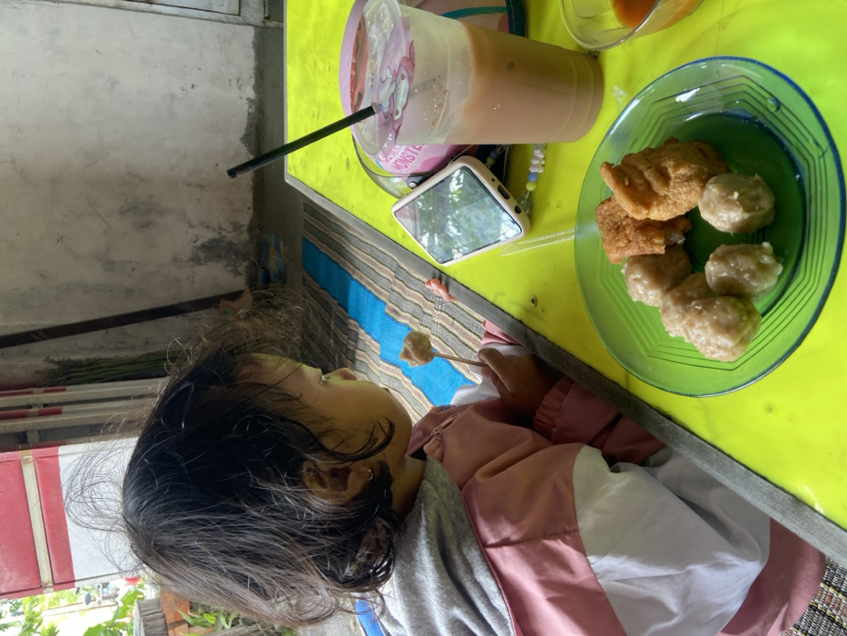 A child with dark hair eats from a skewer at a yellow table. On the table is a clear plastic cup with a straw, a phone, and a green plate of fried tofu and meatballs. The child wears a jacket with pink and white panels. The background includes a gray wall and glimpses of outdoor vegetation. The photo is taken from an angled, overhead perspective.