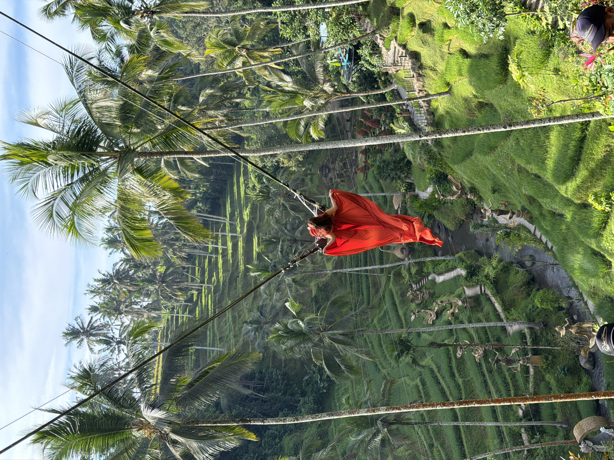 A woman in a flowing red dress is on a swing, suspended by ropes amidst a lush landscape of terraced rice fields and palm trees in Tegallalang, Indonesia. The swing is positioned high above the rice paddies, which are carved into the hillside and appear vibrant green. A narrow stream flows through the valley at the bottom of the terraces. Scattered throughout the rice fields are several stone statues of human figures. The sky is blue with scattered white clouds. The scene is captured from a slightly elevated perspective, looking down into the valley and across the expanse of the rice terraces.