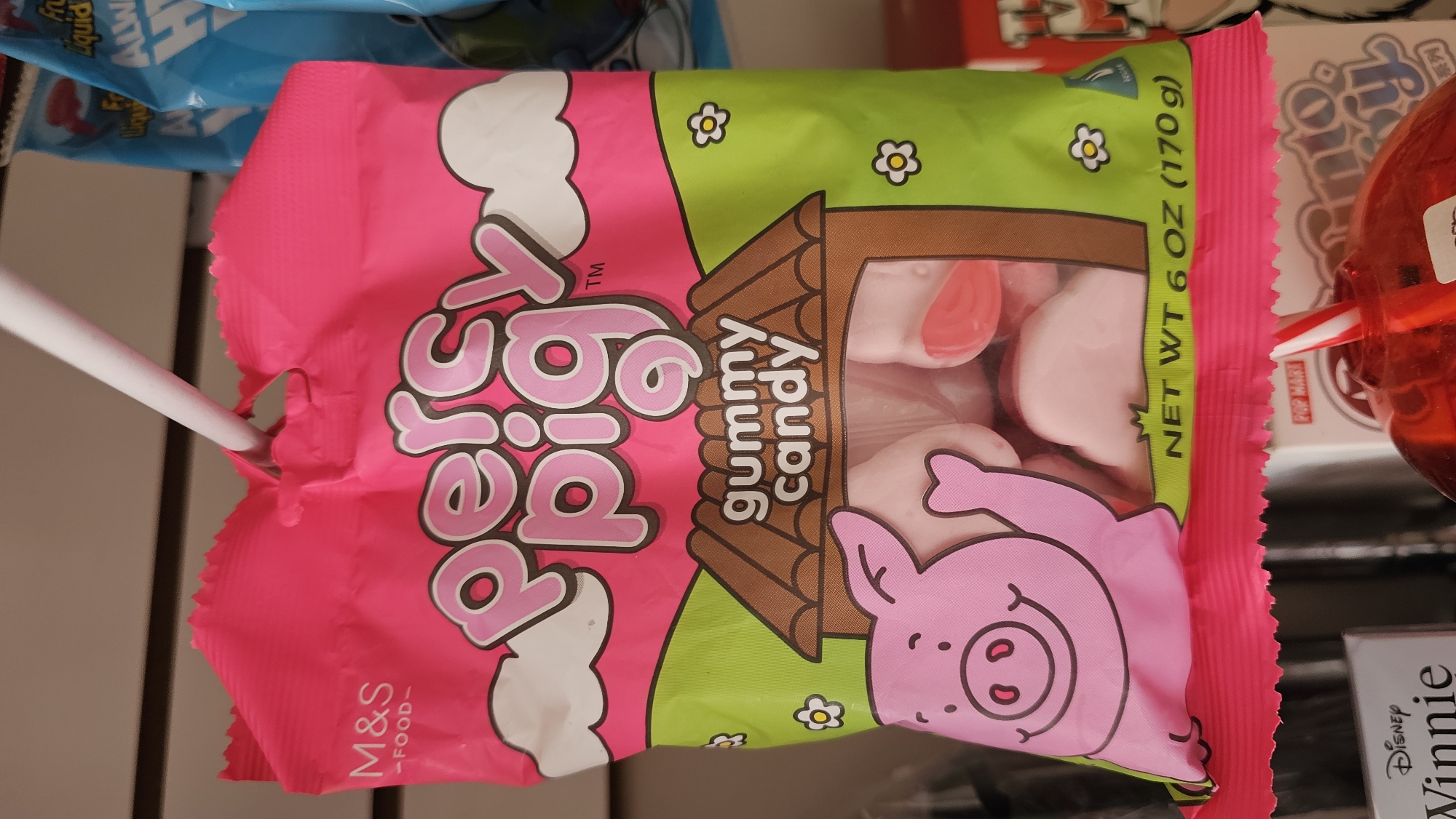 The image displays a bright pink and green bag of "Percy Pig" gummy candy, prominently featured in what appears to be an indoor retail setting, likely in San Bernardino Tlaxcalancingo, Mexico. The candy bag is branded "M&S -FOOD-" and depicts a cheerful pink pig character in a grassy field with a wooden pigsty, white clouds, and yellow flowers. A transparent window on the bag reveals light pink and white pig-shaped gummy candies.
Visible text on the main product includes "Percy Pig" (visually, though partially obscured and stylized), "gummy candy," and "NET WT 6 OZ (170g)." A white rod passes through a punched hole in the top left of the bag, suggesting it is hanging on a display rack.
In the background, to the left, a blue package with partially visible text like "Liquid," "ALW," and "Fr" can be seen. To the right, a red spherical container with a red and white striped straw is visible, along with other merchandise packaging featuring "POP MART," "Disney," and "Winnie." The scene depicts a typical retail display, with various products arranged for sale. No people are present, and the time of day is not discernible.
