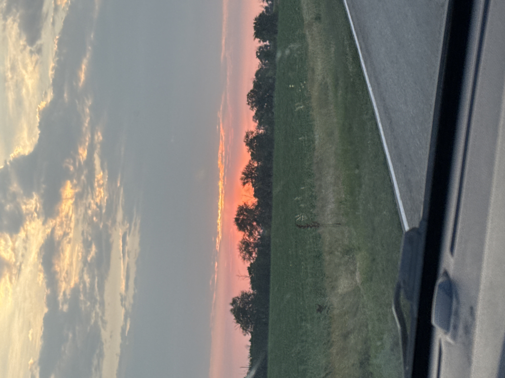 A wide shot captured from inside a vehicle shows a sunset over a rural landscape. The sky is a blend of gray and orange clouds, with the sun visible as a bright orange orb near the horizon. A line of trees forms a dark silhouette against the colorful sky, separating it from a green field below. The field appears to be a cultivated crop, possibly farmland, with some taller grass and weeds visible along the edge. A paved road with a white line marking the shoulder is in the foreground, suggesting the image was taken while driving. The lower portion of the frame shows the vehicle's dashboard and windshield wipers, reinforcing the perspective. The scene is devoid of any human figures or notable structures, emphasizing the natural beauty of the sunset in a tranquil, rural setting. The location is identified as Razvilnoye, Russia.