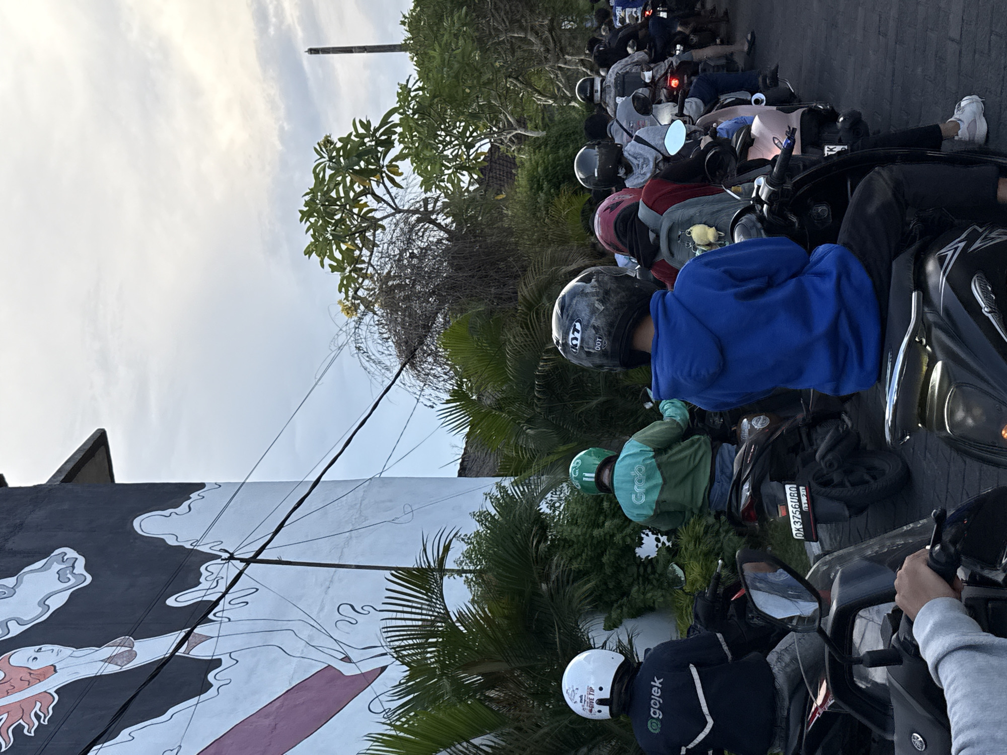 The image shows a street scene in Ungasan, Indonesia. A line of motorcycles is queued on the road, with riders wearing helmets and casual clothing. Some riders are affiliated with ride-hailing services "Gojek" and "Grab" as indicated by their jackets. To the left, a mural is painted on a building, depicting figures and abstract shapes in black and white with hints of red. The surrounding environment includes trees, foliage, and an overcast sky. The license plate number on one of the motorcycles is DK3756UBQ.