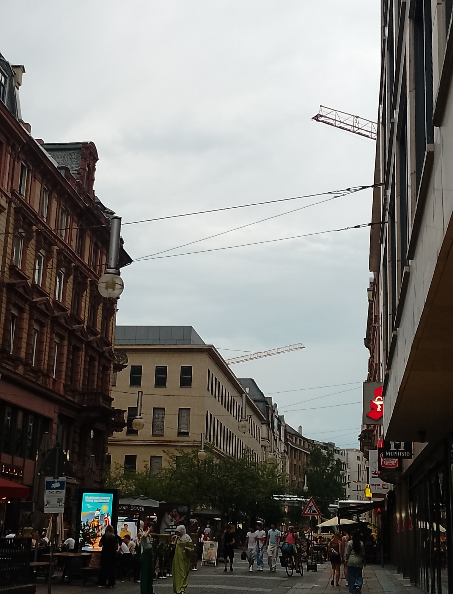 The image shows a street scene in a city. The sky is overcast. On the left, there is a red brick building with classical architectural details, including pilasters and decorative cornices. A street lamp hangs from the building. Further down the street, there are more buildings of varying architectural styles, some appearing to be older and others more modern. One building has a light-colored facade with dark windows. In the distance, a construction crane is visible. On the right side of the street, there are more modern buildings with large windows and signage, including a red pharmacy sign with a caduceus. People are walking on the sidewalk and sitting at outdoor cafes. Some people are dressed casually. The scene suggests a commercial area within a city.