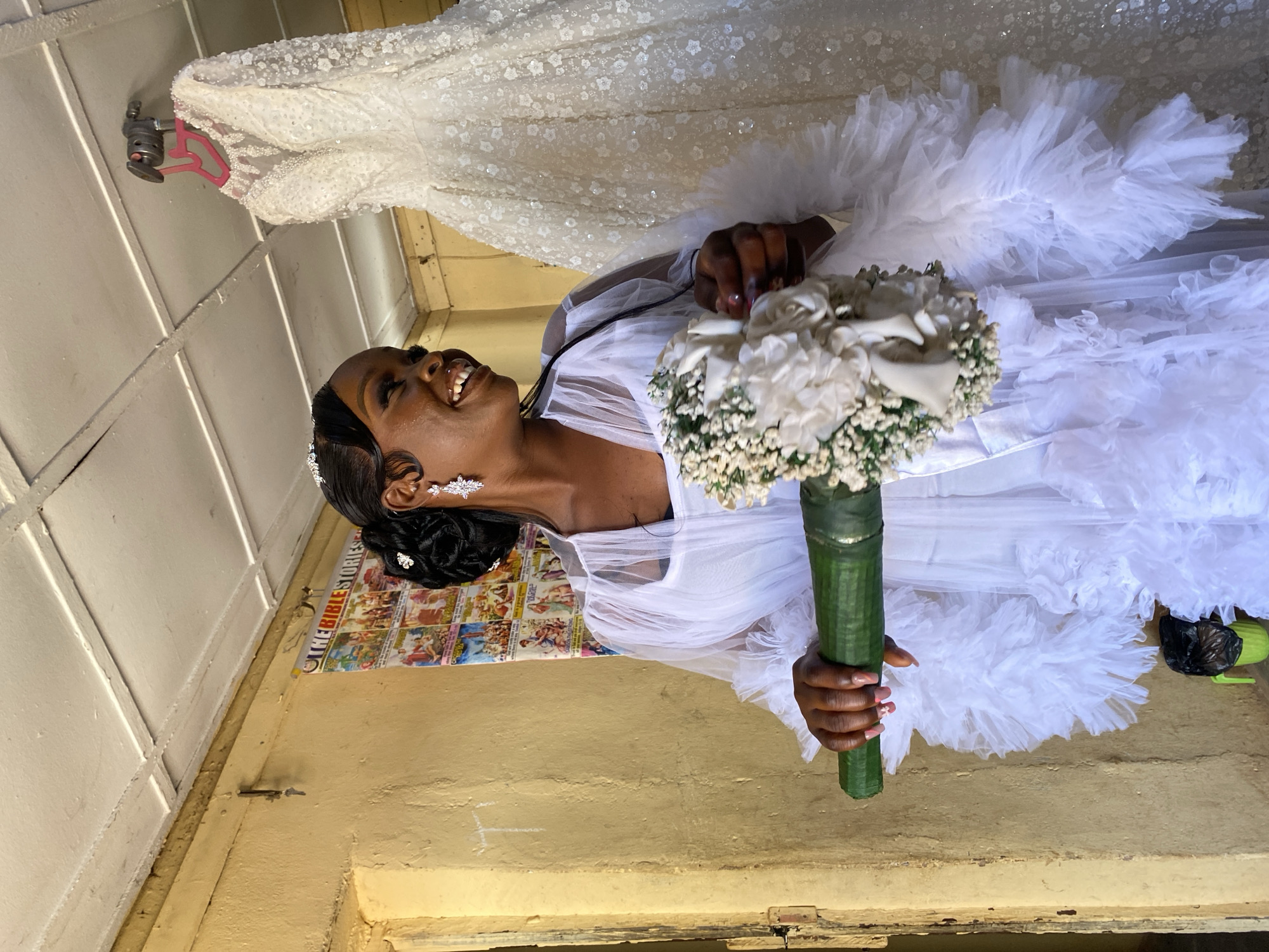 The image depicts a woman in a white wedding dress, holding a bouquet of white flowers with a green stem, in Vom, Nigeria. The woman is smiling and looking upwards. Behind her is a wall with a poster titled "THE BIBLE STORIES" and another white wedding dress hanging above. The room appears to be simply furnished, with a yellow wall.