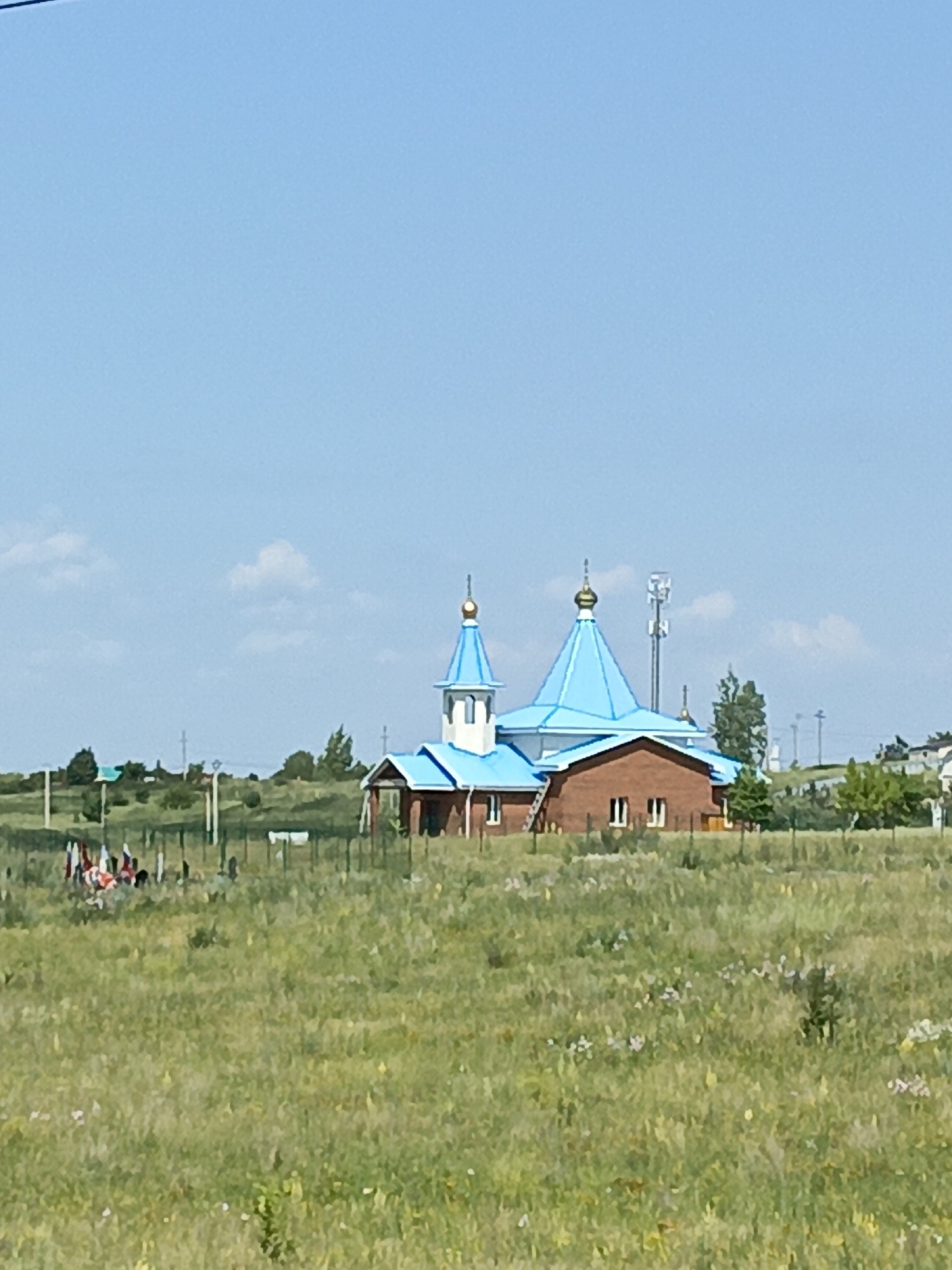 A brick church with bright blue domes stands in a grassy field under a clear blue sky in Lekarevka, Russia. A group of people are gathered in the foreground, some holding flags. The church has a traditional architectural style with multiple domes and a cross on top. In the background, trees and some power lines can be seen, suggesting a rural or semi-rural setting. The field appears to be a meadow with wildflowers.