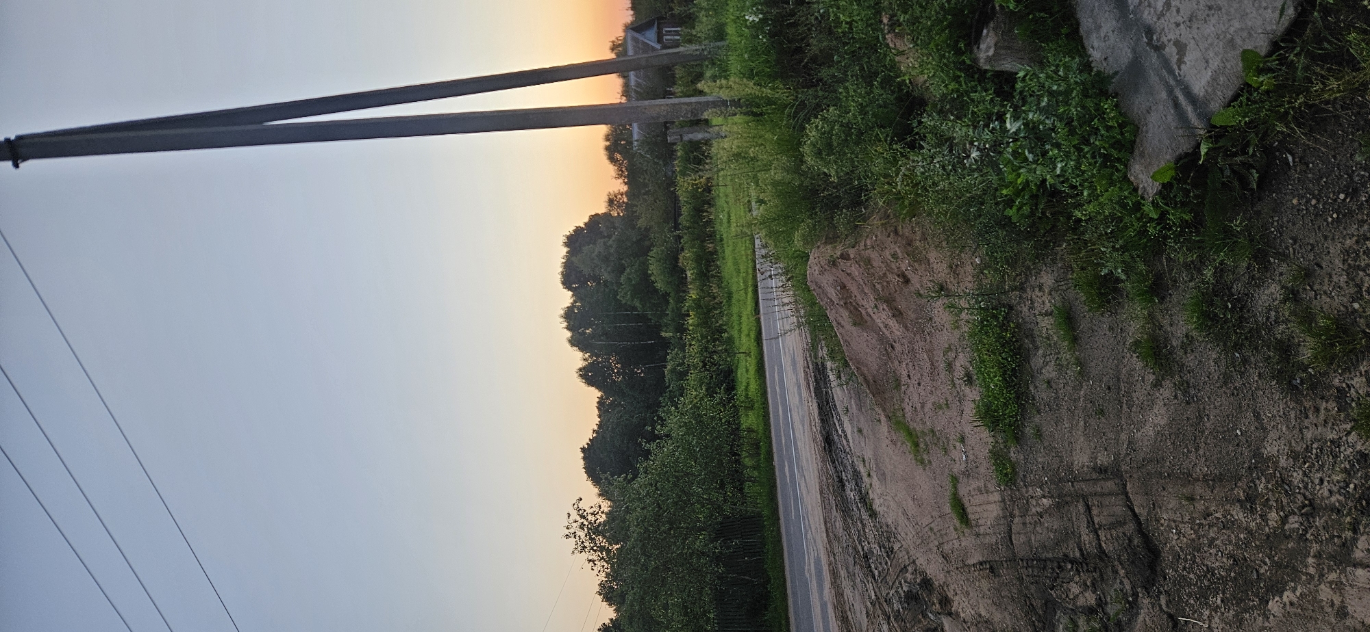 An outdoor vertical medium shot captures a rural landscape at dusk. A light gray sky with a faint orange hue near the horizon suggests the setting sun. In the foreground, a dirt path with tire tracks leads to a paved road. To the right of the path, a pile of reddish-brown dirt sits near dense green foliage and a concrete slab. Two tall, dark gray utility poles stand prominently on the right side of the frame, with power lines stretching across the upper portion of the image. Beyond the road, a thick line of dark green trees forms a backdrop, with a small, light-colored house visible through a gap in the trees on the right. The scene is located in the Mazalawski Selsoviet, Belarus.