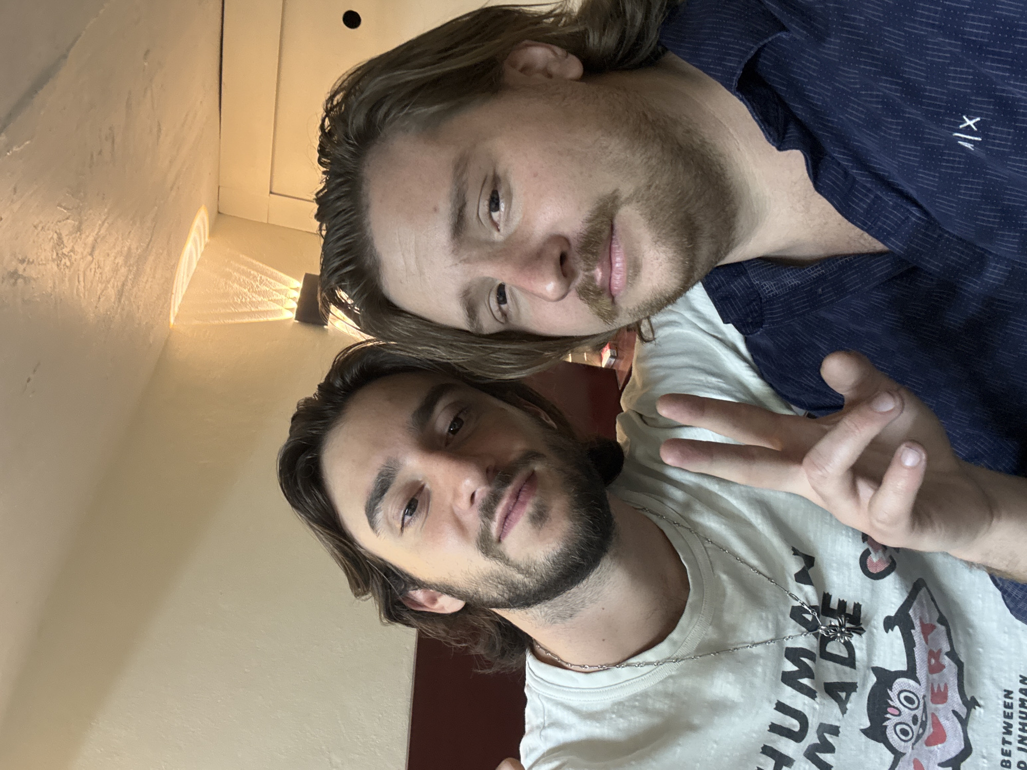 Two men pose closely for a selfie. The man in the foreground wears a white t-shirt with "HUMAN MADE" printed on it, along with a silver necklace featuring a cross pendant, and makes a peace sign with his fingers. The man in the background wears a dark blue patterned shirt. The location is presumed to be indoors, likely a building, given the wall and ceiling visible in the background. There's no information available to confirm that the location is in the city of Huixquilucan de Degollado, Mexico.