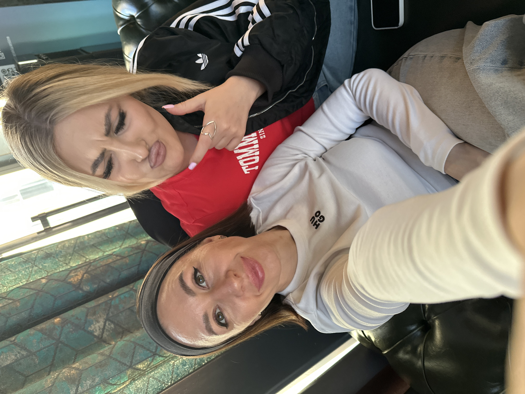 Two women are taking a selfie indoors. The woman on the left, with blonde hair, is wearing a red Tommy Hilfiger shirt under a black Adidas jacket and light blue denim jeans. She is pouting and pointing at the woman on the right. She has a silver heart-shaped ring on her left ring finger. The woman on the right has brown hair with a black headband. She is wearing a long-sleeved white Hugo shirt. They are seated on black leather furniture. There is a table with a patterned surface nearby and a phone lying on the seat. The location is unknown and cannot be inferred from the image.