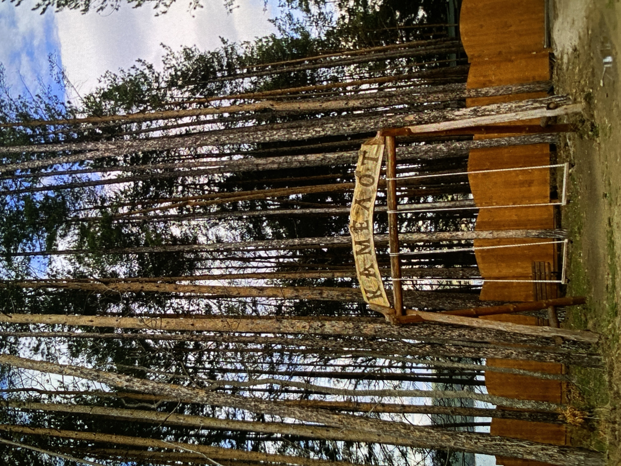 This outdoor image, taken in Bankya, Bulgaria, depicts a rustic recreational area under a partly cloudy sky. The scene is dominated by a unique structure composed of numerous slender tree trunks laid horizontally, forming a textured wall or fence. In front of this log wall, a prominent curved wooden sign, made from a log, displays the word "KAMEЛOT" (Camelot) in carved Cyrillic letters. This sign is supported by two vertical tree posts.
To the right of the sign, a simple wooden swing set stands, featuring a crossbeam from which two swings with white ropes and wooden seats are suspended. Behind the swing set, a dark brown wooden plank fence or wall is visible, possibly part of a nearby structure. The background consists of a dense wooded area with green foliage, suggesting a forest or park setting. The ground in the foreground appears to be a dirt path with some dry grass. The image is captured during daylight hours, conveying a peaceful and natural ambiance, indicative of a place designed for leisure and outdoor activities.