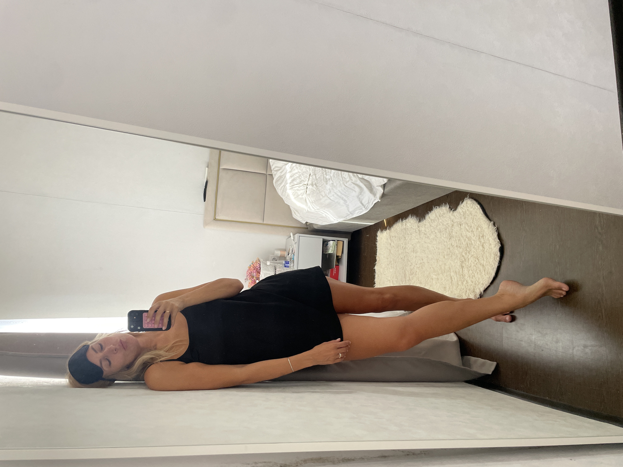An adult female is depicted taking a full-body selfie in a large mirror within a bedroom in Baranovka, Russia. The woman, with blonde hair, stands barefoot, leaning against a light-colored panelled surface that borders the mirror on the left. She wears a black sleeveless mini-dress or nightgown and a black sleep mask positioned on her forehead, obscuring her eyes. She holds a black smartphone, featuring a pink and white textured case, in her right hand. A ring is visible on her left ring finger, and a thin bracelet adorns her left wrist. Her bare legs are slightly crossed, with her left foot pointed.
The reflection in the mirror reveals the bedroom's interior. A bed with a padded, rectangular beige headboard accented with gold trim is prominent, covered by a rumpled white duvet. Adjacent to the bed on the left, a light-colored nightstand holds an assortment of items, including a small basket of colorful objects, various bottles, and a white remote control. The room features dark wood flooring, partially covered by a small, shaggy cream-colored rug situated near the bed. A dark gray curtain is visible to the left of the woman's reflected figure, suggesting a window.