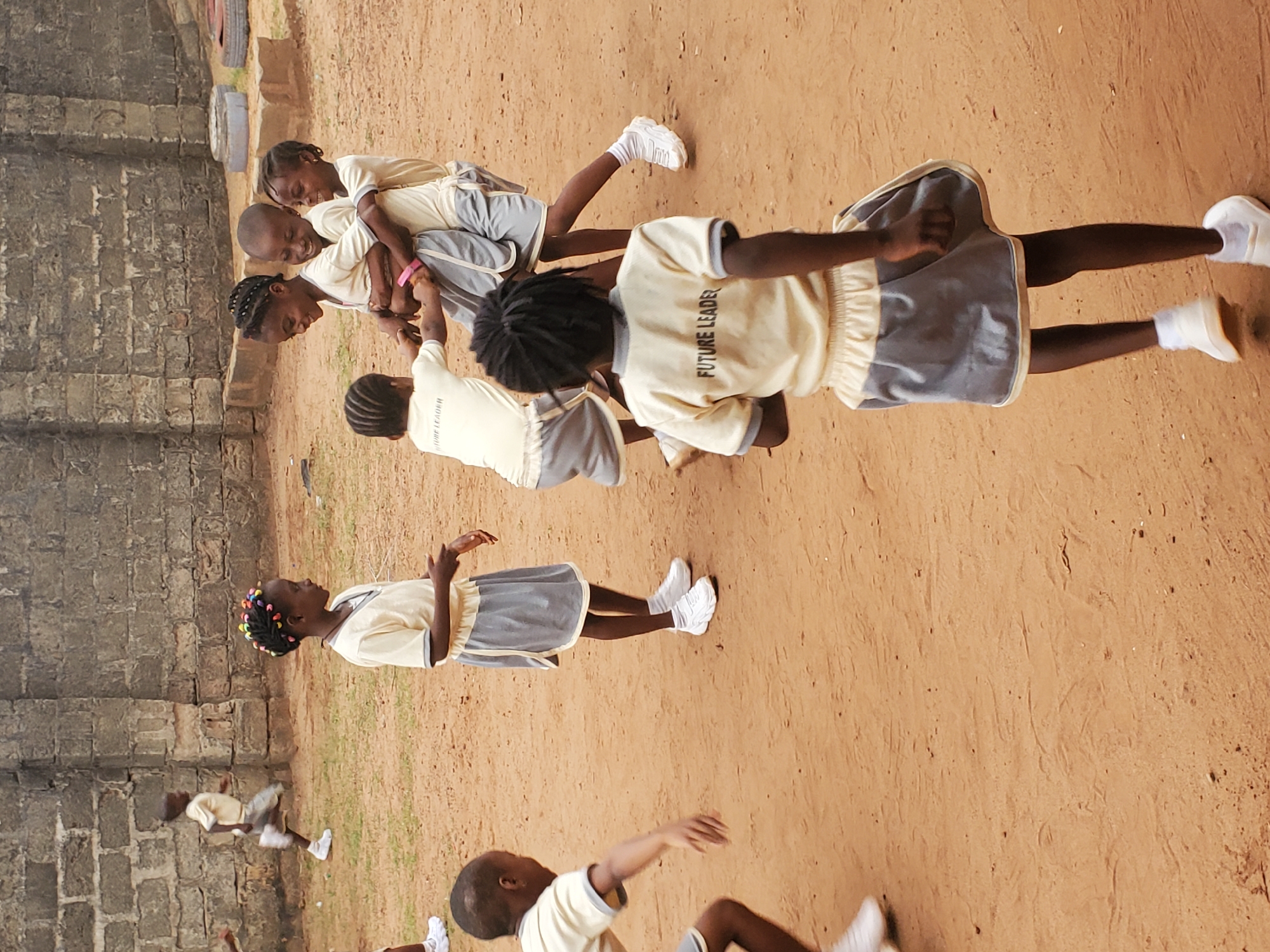 The scene depicts a group of children in a sandy yard in Ukwu-Abwa, Nigeria, likely at a school. The children are wearing matching uniforms, light-colored shirts with "FUTURE LEADER" printed on the back, and grey skirts or shorts, and white sneakers. Several children are involved in a team-building exercise, linking arms. Other children are standing around, some appear to be participating in activities such as running and jumping. In the background is a weathered brick building.