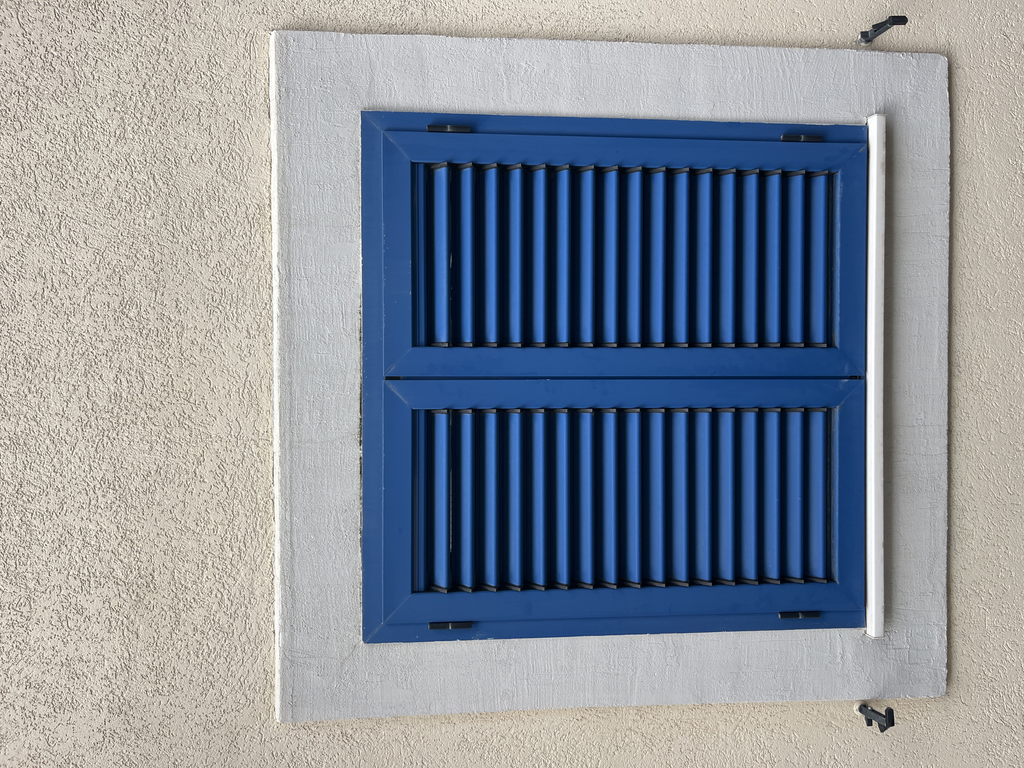 A window with blue louvered shutters is set in a light gray surround, framed by a beige stucco wall. The location is Bečići, Montenegro. A white pipe runs vertically along the right side of the window frame. Above and below the window are black, metal latch components. The shutters are closed.