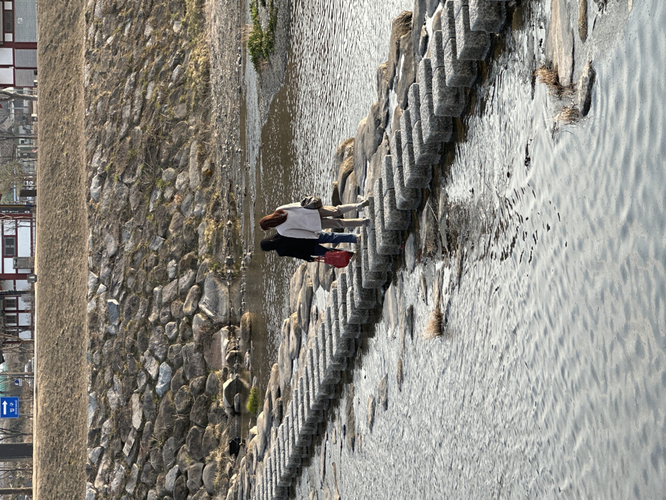 The image depicts a daytime scene at the Muncheon River in Gyeongju-si, South Korea. Two individuals are observed from behind, walking from left to right across a stepping stone bridge composed of regularly spaced rectangular granite blocks. The person on the left wears a dark top and blue pants, carrying a red bag in their left hand. The person on the right wears a light-colored top and light-colored pants, with a dark bag slung over their left shoulder. The river water flows gently around the stepping stones and natural rocks. Beyond the river, a rocky embankment rises, topped with dry, light-brown vegetation. In the background, on higher ground, several traditional Korean-style buildings with red-tiled roofs and white walls are visible. A blue road sign in the upper left background displays white Korean text for "Entrance," "Gyeongju," and "Bomun." The environment suggests an outdoor setting, possibly during a dry season, under clear skies.