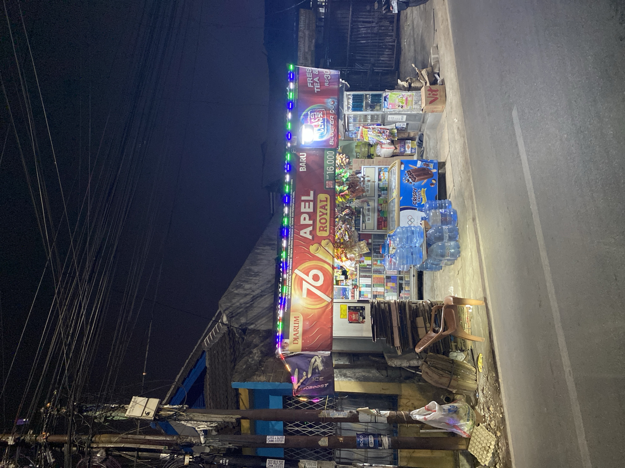 The image depicts a roadside stall or convenience store at night in Tangerang, Indonesia. The stall is illuminated by colorful LED lights above and the glow from within the store. Various products are displayed, including bottled water, snacks, and cigarettes, with prominent signage for "DIARUM 76 APEL ROYAL" and "CLASS MILD BLUETEA DUO". In front of the store, several large bottles of drinking water are stacked. To the left of the store, there is a pile of cardboard boxes and a single plastic chair. A motorcycle is partially visible on the right side of the image. The environment suggests an urban setting with a street in the foreground and electrical wires overhead. There are no people visible in the image.