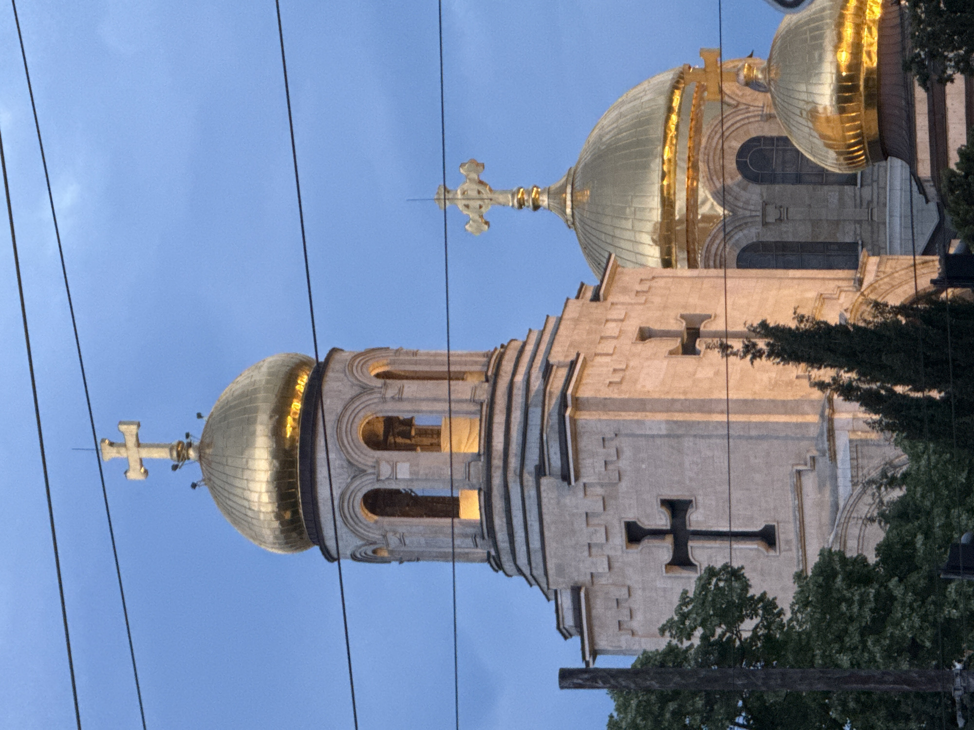 The image depicts the exterior of the Dormition of the Mother of God Cathedral in Varna, Bulgaria. The view is from below, looking upward at the cathedral's domes and towers. Two prominent gold-colored domes are visible, each topped with a cross. The stone exterior of the cathedral features carved cross details. Overhead power lines run diagonally across the sky. Trees are visible at the lower right of the frame. The sky is a light blue.