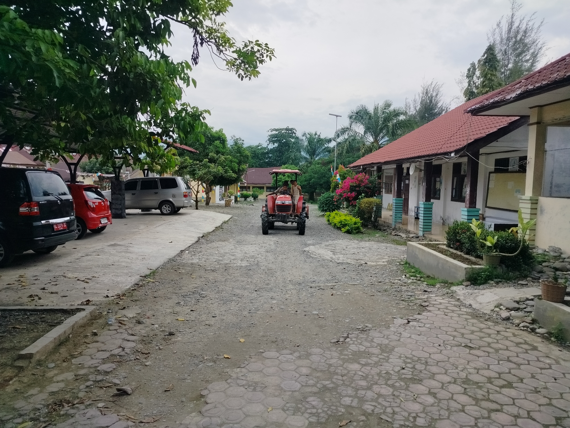 The image depicts a view of a street in Purwodadi, Indonesia, under an overcast sky. A red tractor with two occupants is moving away from the camera down a gravel road. On the left side of the road, a black SUV and a red car are parked. Beyond the parked cars, there is a building with a red-tiled roof, partially obscured by trees. On the right side of the road, another building with a red-tiled roof is visible, extending further into the distance. The buildings appear to be low-rise structures with white walls. Green trees and bushes are present throughout the scene. The ground is a mix of gravel and paved sections with a hexagonal pattern.