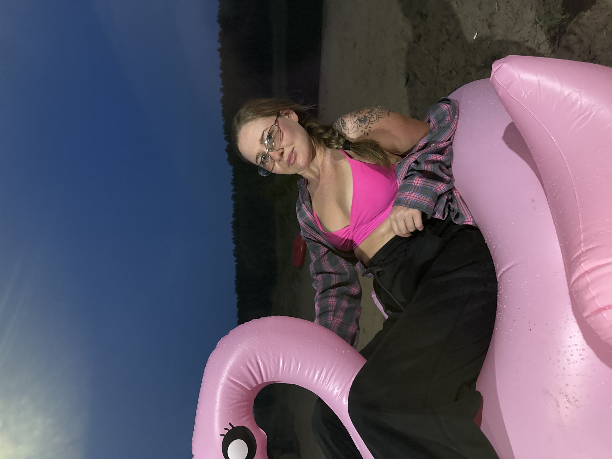 A woman with blonde hair in a braid and glasses, wearing a pink bikini top and a plaid shirt, reclines on a pink inflatable flamingo on a sandy beach. A dark blue sky with a hint of a sunset or twilight is visible in the background, along with a line of trees bordering the water. The scene appears to be set in the evening. The image does not provide any specific information or visual cues to identify the location as Garevaya, Russia.