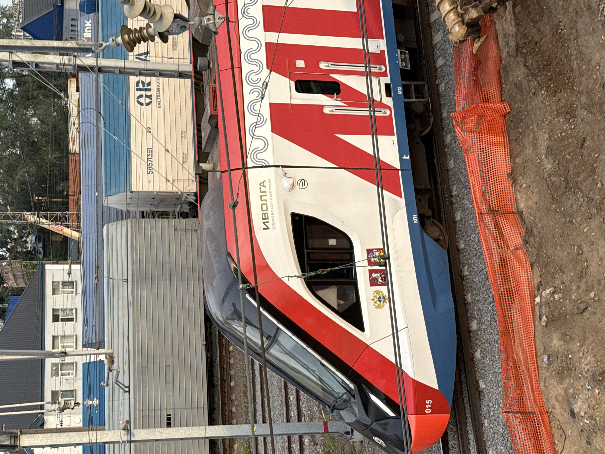 The image shows a modern commuter train, identified as "ИВОЛГА" (Ivolga), with its serial number "015" visible on the front. The train is white with red and blue accents, featuring a stylized wave pattern and large red letters that appear to spell out "МУ". Emblems of the Russian Federation are also visible on the side of the train. The train is on railway tracks, with overhead power lines characteristic of an electrified railway system.
In the background, there are several other trains or railcars, some appearing to be cargo or freight cars, with one displaying the text "RAILTRANS AUTO" and a serial number. Industrial buildings, possibly part of a train depot or a logistics facility, are also visible. The environment suggests an urban or industrial setting within a city. The presence of construction netting and disturbed ground in the foreground indicates ongoing work or maintenance on or near the railway.
This train, the Ivolga, operates on Moscow Central Diameters (MCD), a suburban railway line in Moscow, Russia. The context of multiple railcars and infrastructure points to a location at a train station or a railyard within the Moscow region.