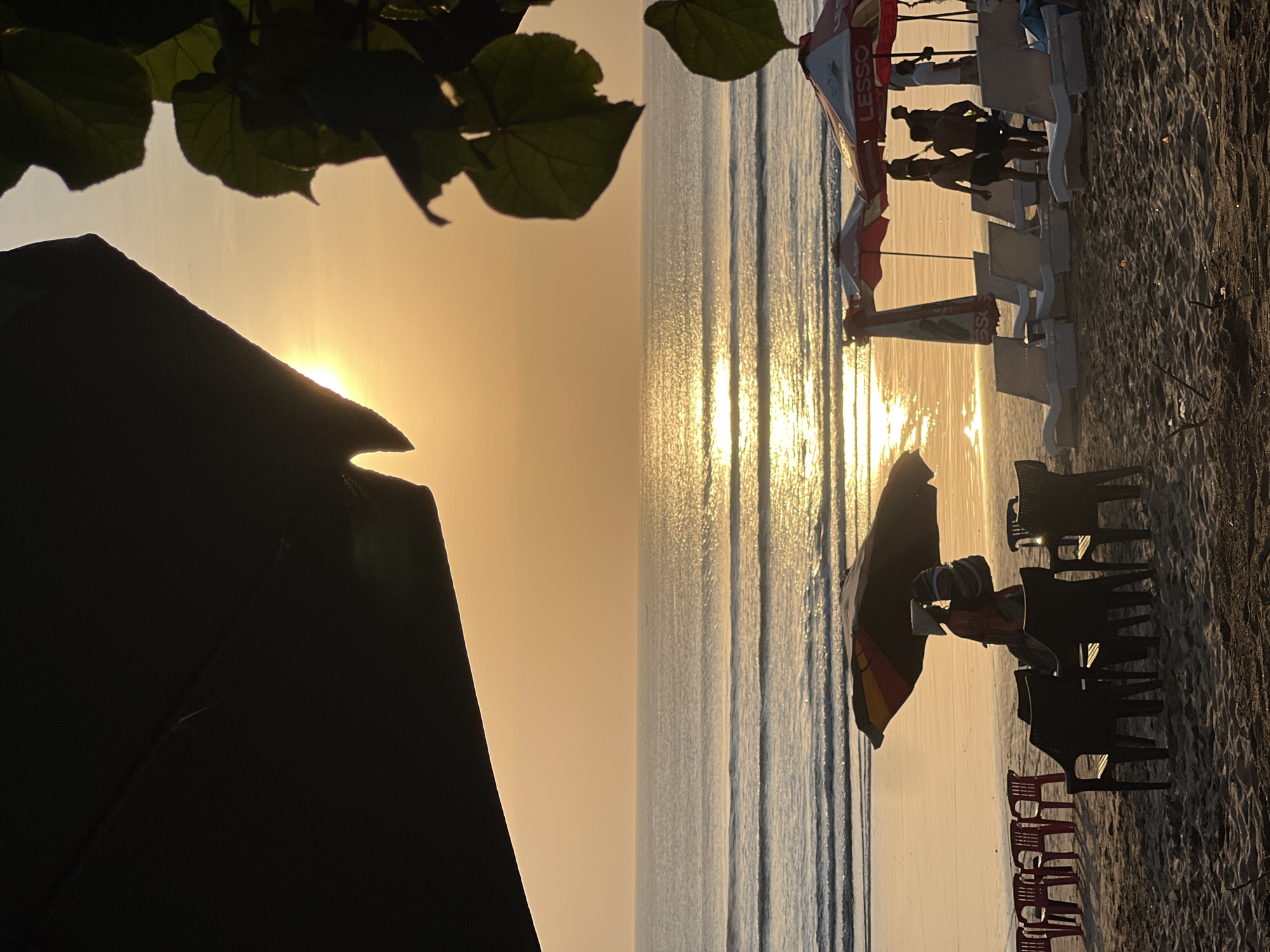 This image depicts a tranquil sunset scene on Kuta Beach, Indonesia. The sun is setting over the ocean, casting a warm golden glow on the water and the sand. Waves gently roll towards the shore, creating a peaceful atmosphere.
In the foreground, lounge chairs and umbrellas are set up on the beach, suggesting a tourist area. Several people are visible, some relaxing on the chairs while others are walking along the shore. The silhouettes of these individuals are softened by the setting sun.
The environment is a typical beach setting with sand, sea, and sky. The presence of beach chairs and umbrellas indicates that this is a popular spot for visitors. The overall mood of the image is one of serenity and relaxation, capturing the beauty of a sunset on a tropical beach.