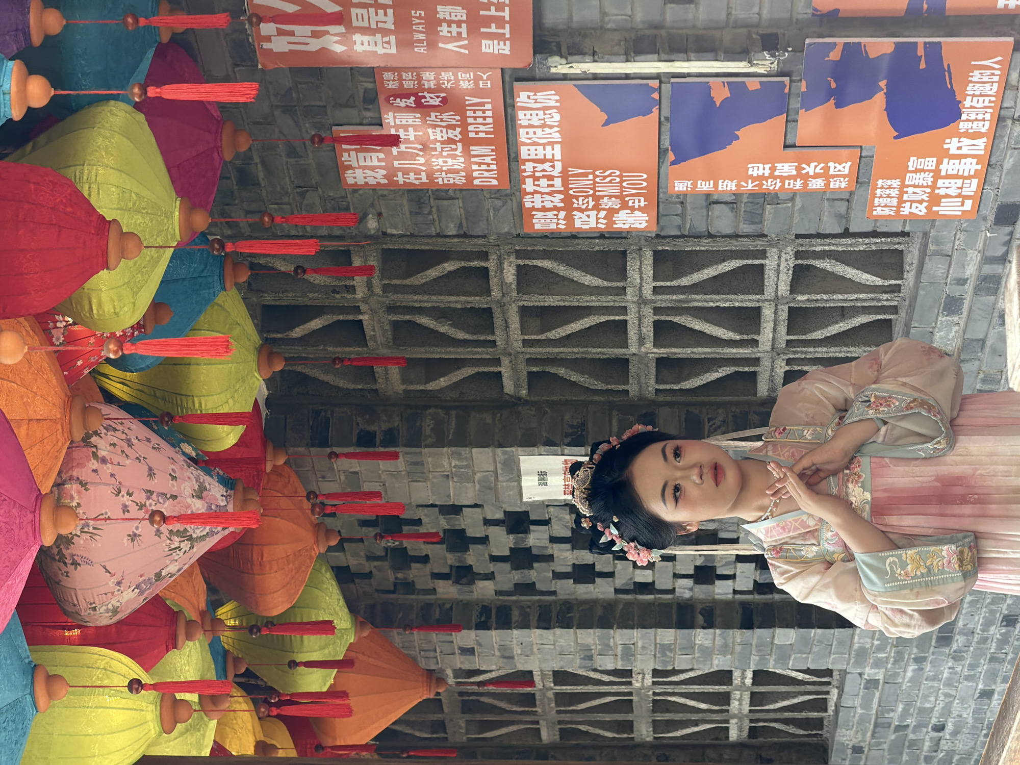 The image shows a young woman dressed in traditional Chinese clothing standing in front of a brick wall. She has her hands clasped in front of her chest and is looking to her right. The wall is decorated with several orange signs with Chinese characters and some English text. Above the wall, a variety of colorful lanterns are hanging, some of which have red tassels. The architectural style and the content of the signs suggest the location is likely Yuzhong District, Chongqing, China, a city known for its blend of historical sites and modern developments. The scene appears to be set outdoors, possibly in a cultural or tourist area.