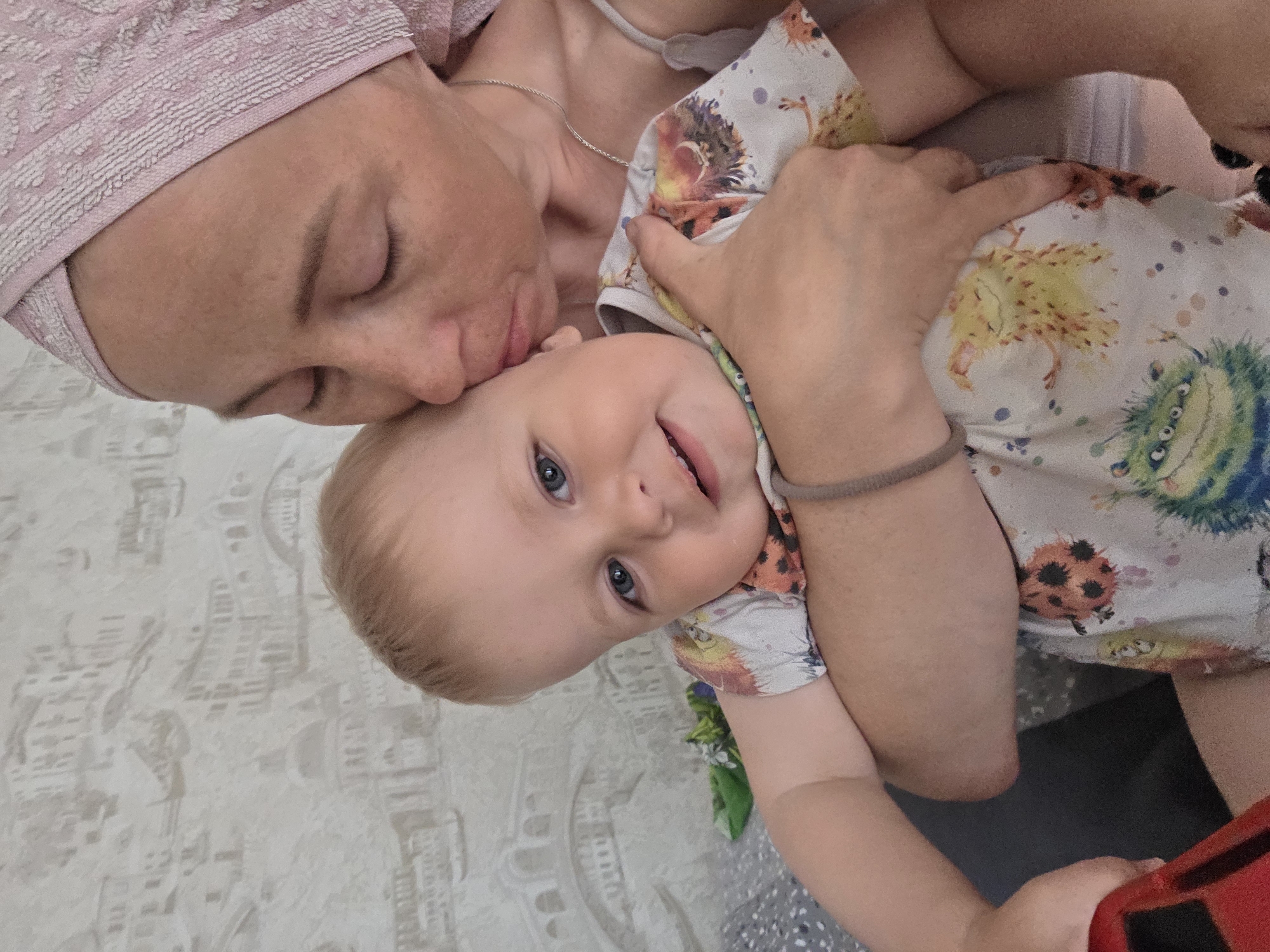 A woman with a pink head wrap is holding a baby in her arms, with her head tilted and eyes closed as if kissing the baby's forehead. The baby is looking at the camera with a smile. The baby is wearing a light-colored shirt with cartoon monster prints. The background is a patterned wall, and there is an arm with a bracelet holding the baby. The image appears to be taken indoors, with an unknown location.
