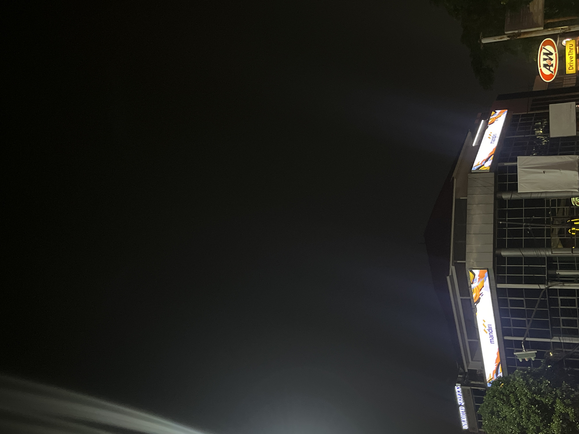 The image shows a nighttime street view in West Jakarta, Indonesia. A multi-story building with glass windows is illuminated by various signs. A prominent horizontal sign on the building displays the word "mandiri" with an abstract colorful design above it. To the right of this building, an A&W restaurant sign with "Drive Thru" is visible. A traffic light is positioned in front of the building. A large tree with dark green foliage is on the left side of the frame, obscuring part of the building. The sky is dark and cloudy, indicating a night scene. The overall environment appears to be an urban commercial area.