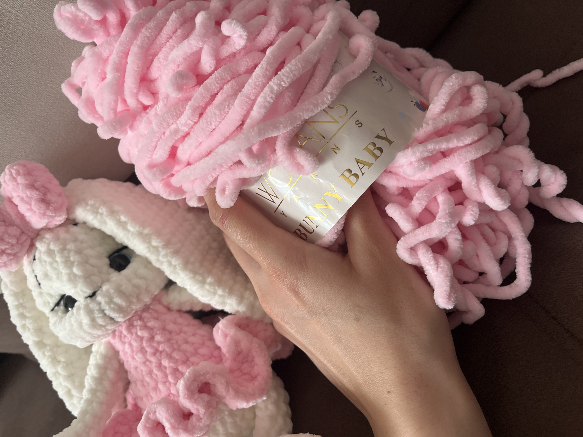 The image shows a skein of pink yarn labeled "Wollans Bunny Baby" held in a person's hand. To the left of the yarn is a crocheted white and pink bunny plush toy. The background is a brown, possibly upholstered surface. The setting is unknown, and there is no evidence to connect the image to Pernik, Bulgaria.