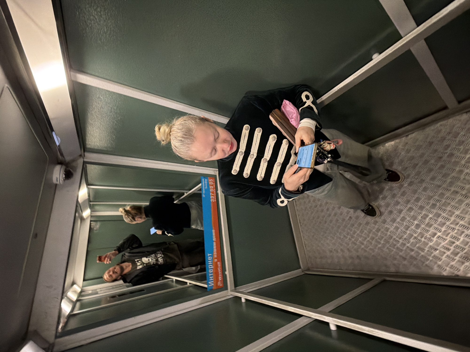 Here is a detailed description of the image:
**Setting:**
The image appears to be taken inside a small elevator or lift with a metallic floor and green walls. The lighting is somewhat dim.
**People:**
- There is a woman who seems to be the main subject of the image. She has blonde hair styled in a bun and is wearing a dark military-style jacket with light-colored embellishments on the front. She is holding a phone and some other items in her hands.
- The reflection of other people is visible in the mirror; a man wearing a black hoodie and a blonde woman who's also using her phone.
**Objects:**
- A mobile phone held by the woman in the foreground.
- A pink bag and what appear to be wooden objects (possibly make-up brushes) in the woman's hands.
- Internet services advertisement.
**Location:**
The sign advertises "WestCall" internet services, telephone and IP-television with the telephone number "337 50 05".
Based on this information and the company advertised, it is plausible that this image was taken in the city of **Saint Petersburg, Russia**, where WestCall is known to operate.