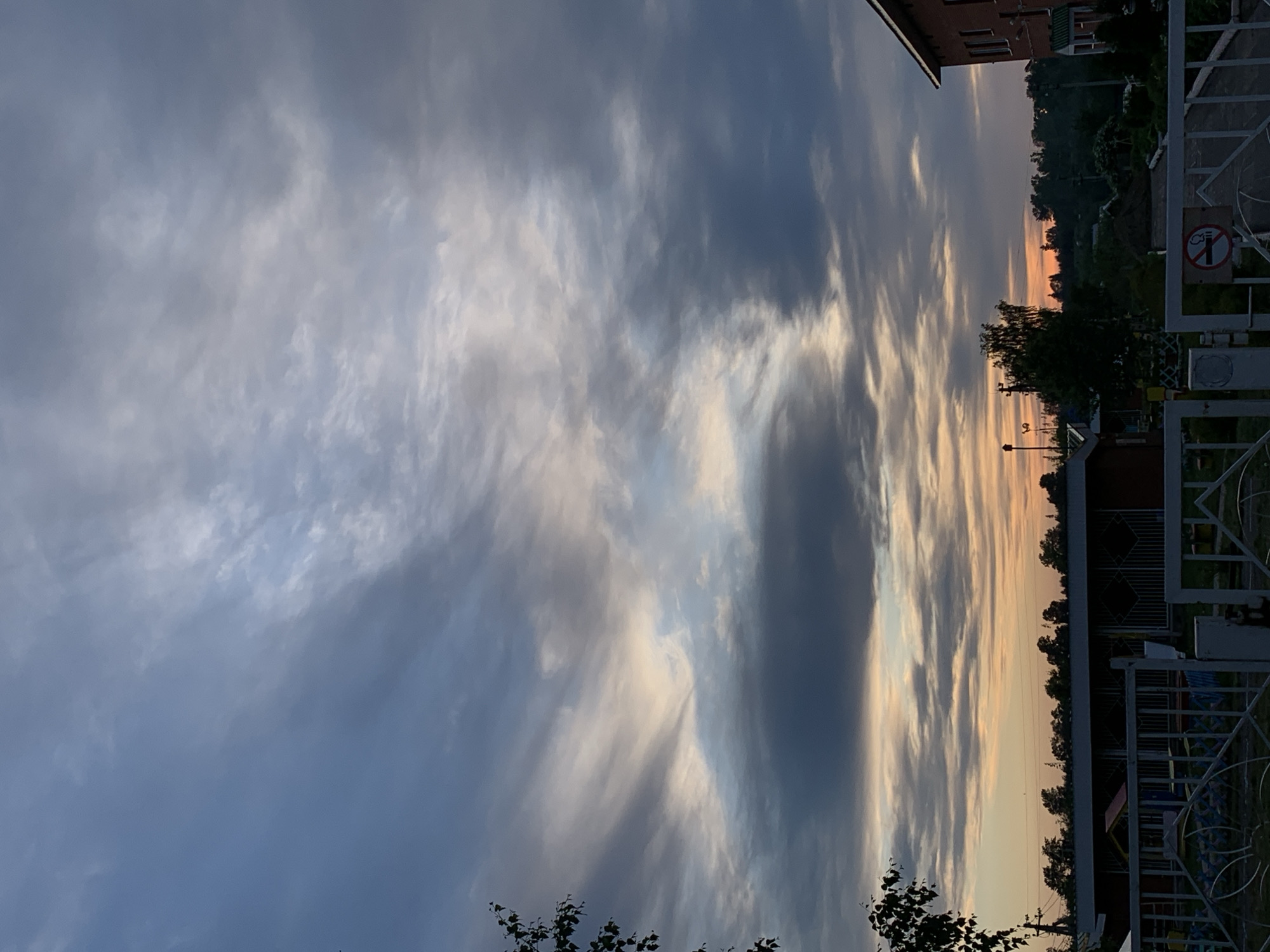 The image captures a sunset scene in Фоки, Russia. The sky is filled with layered clouds illuminated by the setting sun, transitioning from blue-gray hues to streaks of orange and yellow near the horizon. The foreground contains trees and urban infrastructure, including fences and buildings. There are no people visible in the image. A no-smoking sign is visible on the fence.