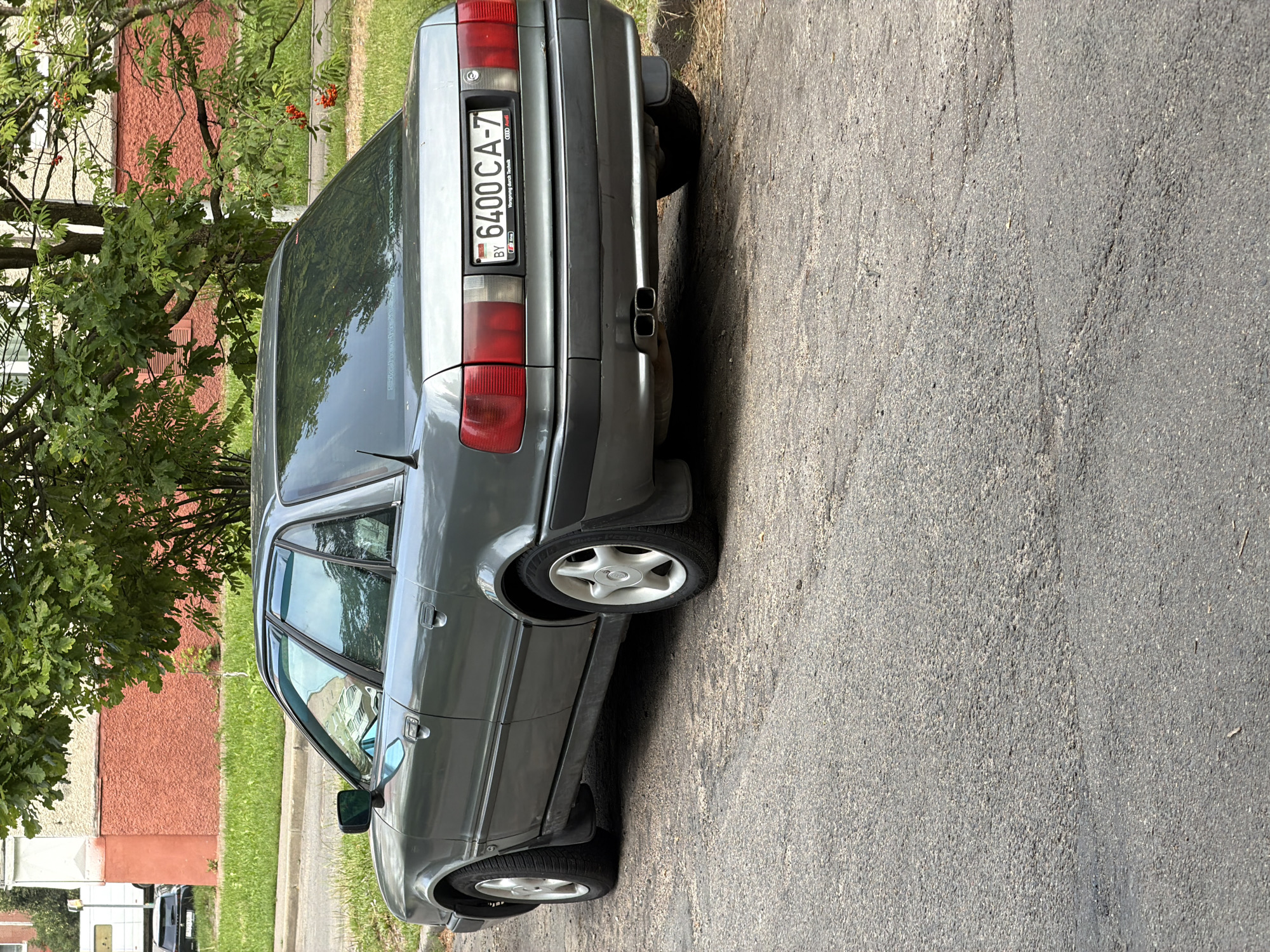 The image shows the rear passenger side of a gray Audi 100 C3 sedan parked on an asphalt road in Minsk, Belarus. The car has a Belarusian license plate reading "6400 CA-7". The background includes a red building with a textured facade, a sidewalk, green grass, and leafy trees with red berries. The day appears to be overcast or the lighting is diffused. There are no people or other vehicles clearly visible in the immediate vicinity of the car.