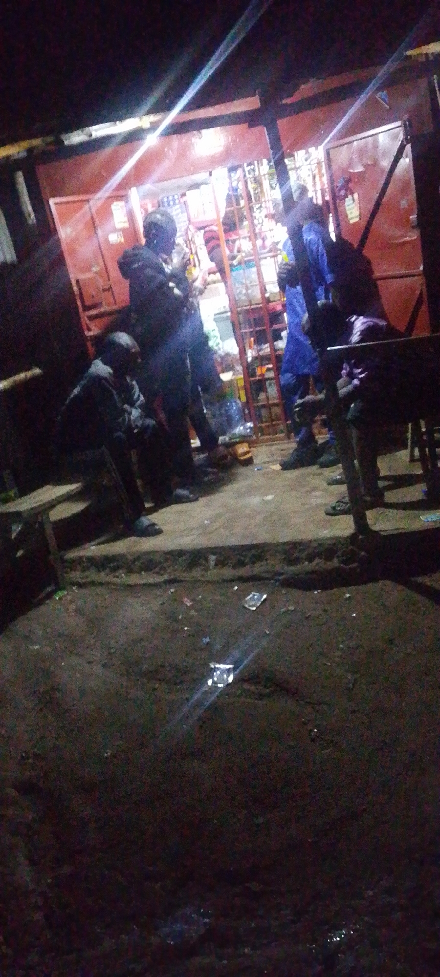 In Gura Top, Nigeria, a nighttime scene unfolds outside a small, open-fronted shop. The shop is dimly lit from within, revealing shelves stocked with various items. A red-painted structure forms the shop's facade, with a metal grille or gate partially visible. Several individuals are present. One person, wearing a dark jacket and pants, is standing near the entrance. Another individual in a blue outfit stands to the right, and a third person, in a dark jacket, is seated on a wooden bench in the foreground to the left. Further to the right, another person is seated, partially obscured by shadow and what appears to be furniture. The ground in front of the shop is dirt, with scattered debris, including plastic wrappers and possibly a water bottle. The overall atmosphere suggests a casual gathering or interaction outside a local business at night.