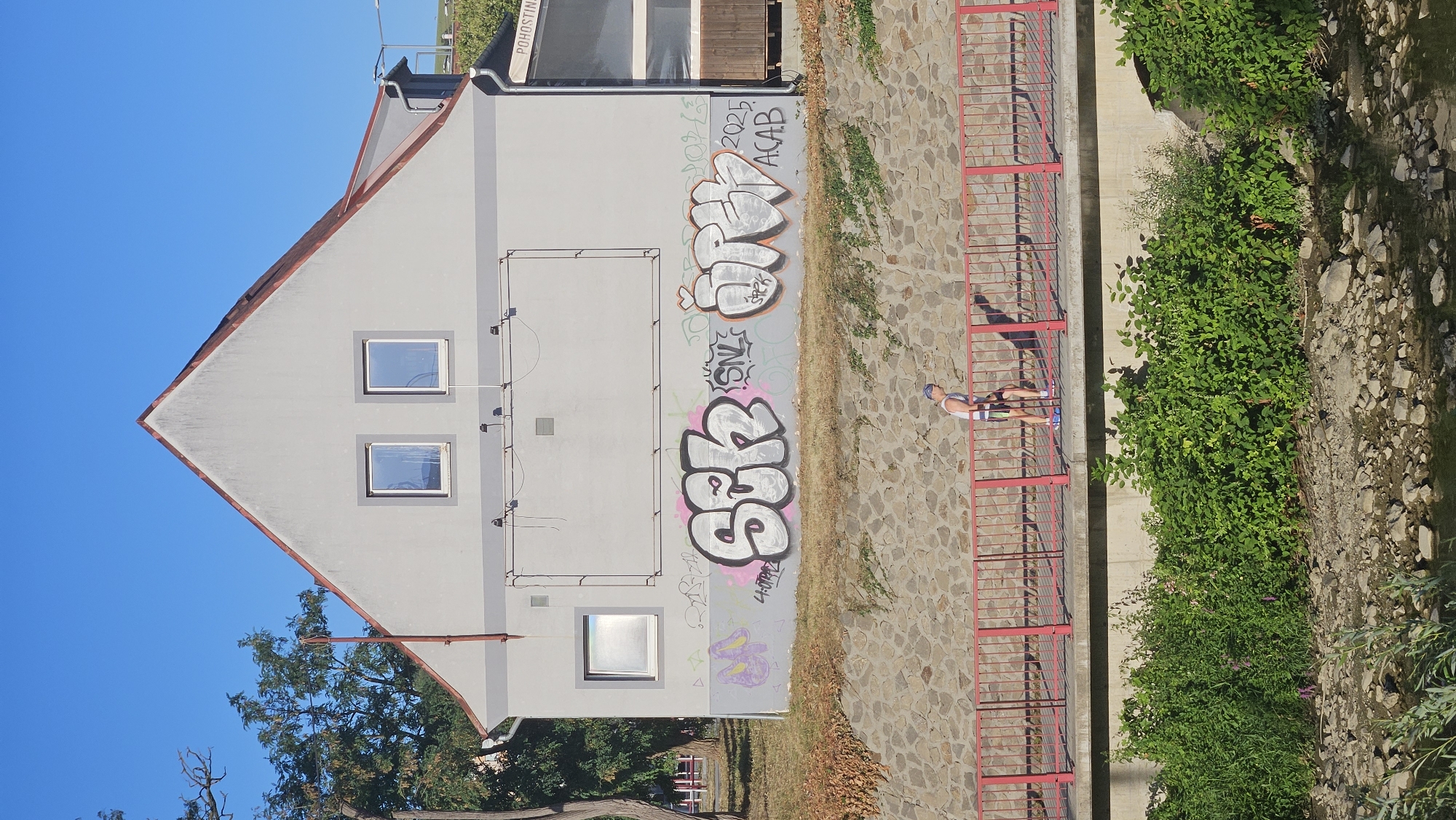 The image shows a building with graffiti on its lower section. The building has a light grey facade, a steeply pitched roof, and two square windows on the upper story. Below these windows, there is a large blank rectangular area that appears to be a sign or billboard space, framed by a thin grey line. Two smaller square windows are visible on the lower portion of the building, one on the left and one on the right.
Graffiti in large, bubble-style lettering covers the lower part of the building's facade. The most prominent graffiti reads "SER" in white with pink outlines, and to its right, a similar style graffiti reads "JRN" in white with orange outlines. Other smaller tags and markings are also present.
In front of the building, a paved walkway runs alongside a low, stone-faced retaining wall. A red metal railing lines the edge of the walkway. A person, appearing to be a male wearing a white tank top and dark shorts, is walking on the walkway, facing left. They are wearing a blue bandana and blue sneakers, and appear to be either jogging or walking briskly. The person casts a distinct shadow on the walkway due to the sunlight.
The area beneath the walkway is overgrown with dense green vegetation. Below the vegetation, the shallow water of a stream or river is visible, with a rocky bed.
The building is located on the right bank of a river or stream, which is visible in the foreground. The overall impression is an urban or semi-urban environment, possibly a recreational area or a commercial building adjacent to a waterway. The building features a sign that reads "POHOS" followed by obscured text, suggesting it might be a restaurant or a similar establishment. The presence of graffiti indicates an area with street art culture. Based on the context of similar imagery and the architectural style, this location is identified as being along the Sekčov riverbank in Prešov, Slovakia, specifically near the bus station area.