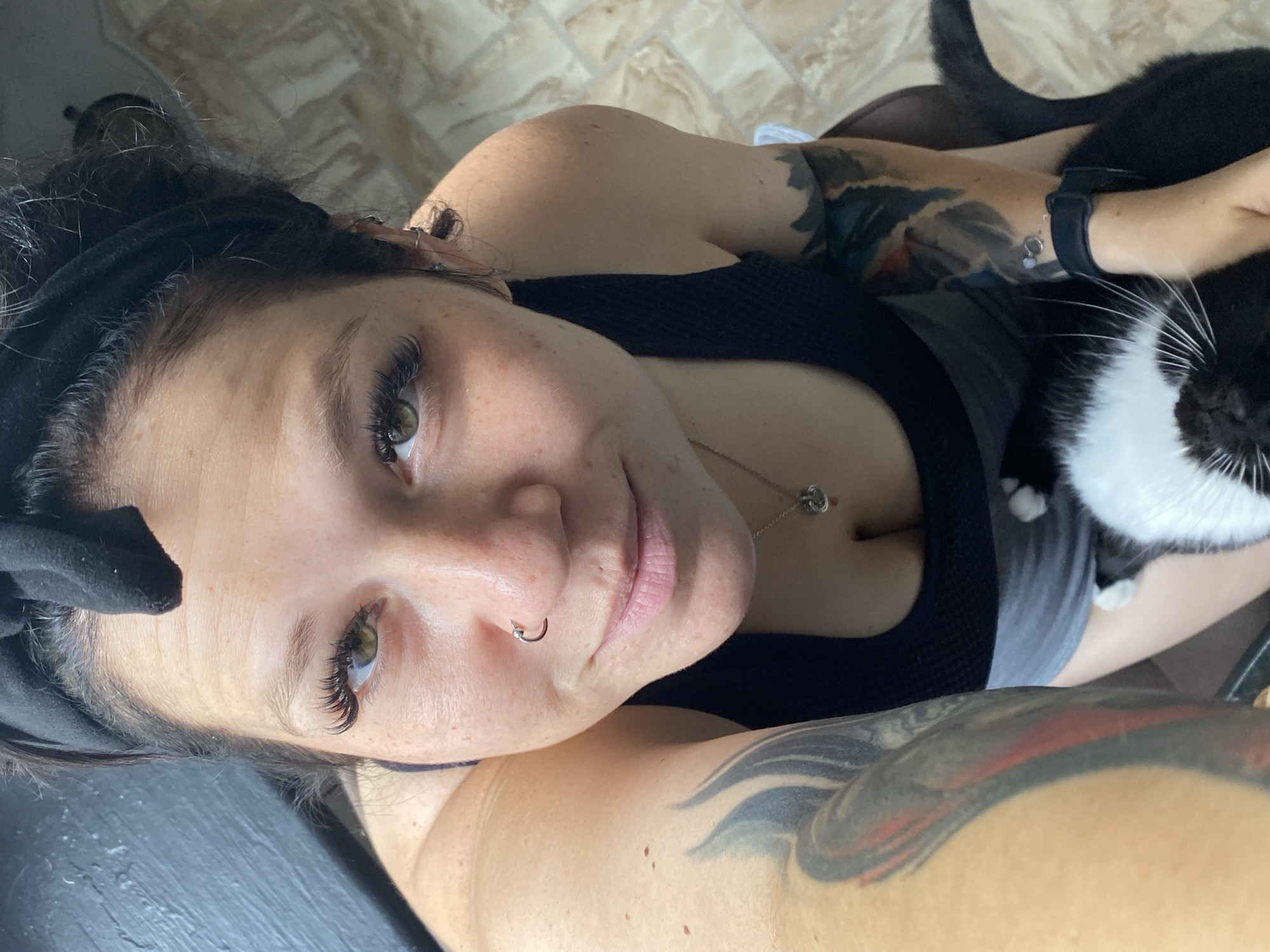 A high-angle, close-up, indoor selfie shows a Caucasian woman with light skin, dark hair pulled back in a black headband, and visible freckles. She is wearing a black tank top and grey shorts. She has voluminous false eyelashes, a nose ring, and large hoop earrings. She has extensive tattoos on her arms. She is holding a black and white cat in her arms. The background appears to be a tiled floor and a dark wall. There is no discernible location in Chaikovsky, Russia, evident in the image.