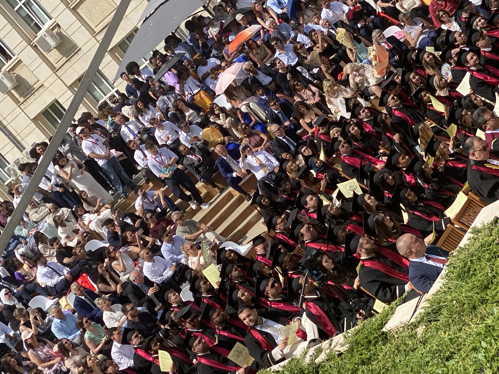 The image shows a large outdoor gathering, likely a graduation ceremony, with attendees seated in rows. Some are wearing academic regalia (black gowns with red stoles), suggesting they are graduates. Other attendees appear to be guests, dressed in a variety of casual and formal attire. Some are using umbrellas for shade. The event takes place in an outdoor venue with a building in the background. The location cannot be verified as Pleven, Bulgaria.
