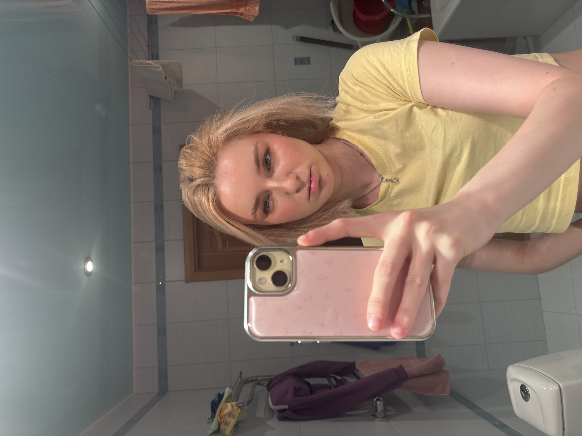 A young woman with blonde hair, wearing a yellow t-shirt and a necklace, is taking a selfie in a bathroom mirror. She is holding a pink smartphone with a floral pattern. The bathroom has tiled walls, a light blue ceiling with a spotlight, a toilet, and a towel rack with clothes. The woman has makeup on her face, including blush and eyeshadow.