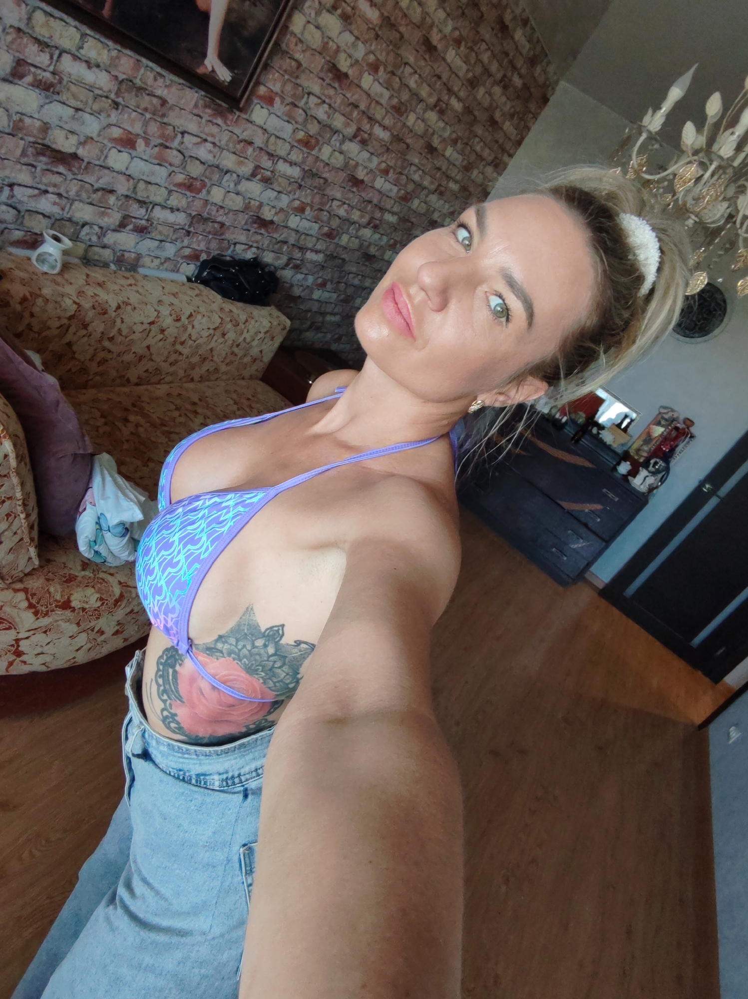 A woman with blonde hair pulled back in a ponytail, wearing a light purple bikini top and light blue denim jeans, poses for a selfie in what appears to be a residential setting. She has a large rose tattoo on her left side. The background includes a brick wall, a patterned couch, a dark dresser, and a chandelier. The flooring is wood.