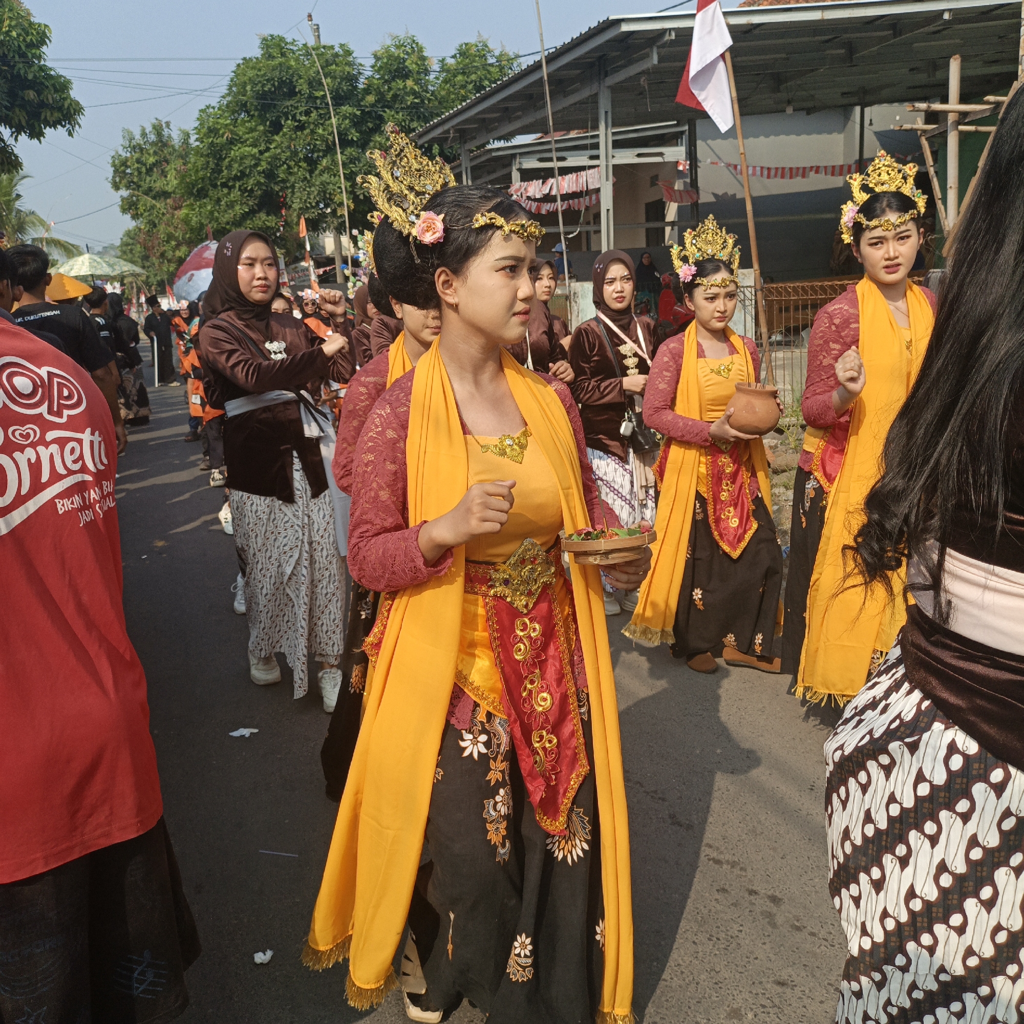 This is a medium shot, eye-level, outdoor, daytime, full-color photograph taken in Tanahbaya, Indonesia. The scene depicts a procession or parade taking place on a paved street lined with trees and buildings. Several individuals, primarily young women, are featured prominently.
In the foreground, a young woman in the center is dressed in traditional attire, including a gold crown, a dark maroon lace top with a yellow sash draped over her shoulders and flowing to the ground. Her skirt is black with white floral embroidery. She is holding a small, woven circular tray with some items on it. To her left, another young woman in similar traditional dress, though her top is dark maroon and her skirt is a lighter brown with a patterned design, is holding a small terracotta pot.
Behind these two central figures, a procession of other individuals in dark maroon tops and patterned skirts are visible, some wearing headscarves. To the far left, a person in a red shirt with white lettering, "ORINETTI" prominently displayed, is partially visible from the back. Other participants and spectators are seen in the background, with umbrellas and flags also present. The environment suggests a warm, sunny day with a clear blue sky. The overall atmosphere appears to be festive and cultural.