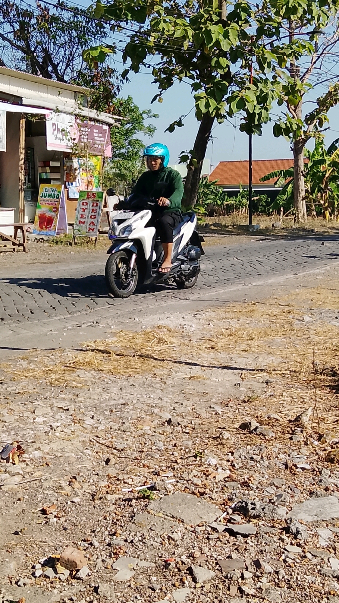 A person wearing a blue helmet and a green hooded jacket rides a white and silver scooter on a paved road. The scooter has a license plate that is not legible. The person is wearing black pants and flip-flops. The road is paved with cobblestones and is dusty. To the left of the road is a small building with a white facade and a wooden counter. Signs in Indonesian are displayed on the building, including one that says "Es Teh Jumbo." There are trees with green leaves and a clear blue sky in the background. A red-tiled roof of a house is visible in the distance. The ground around the road is covered with dry grass and debris.