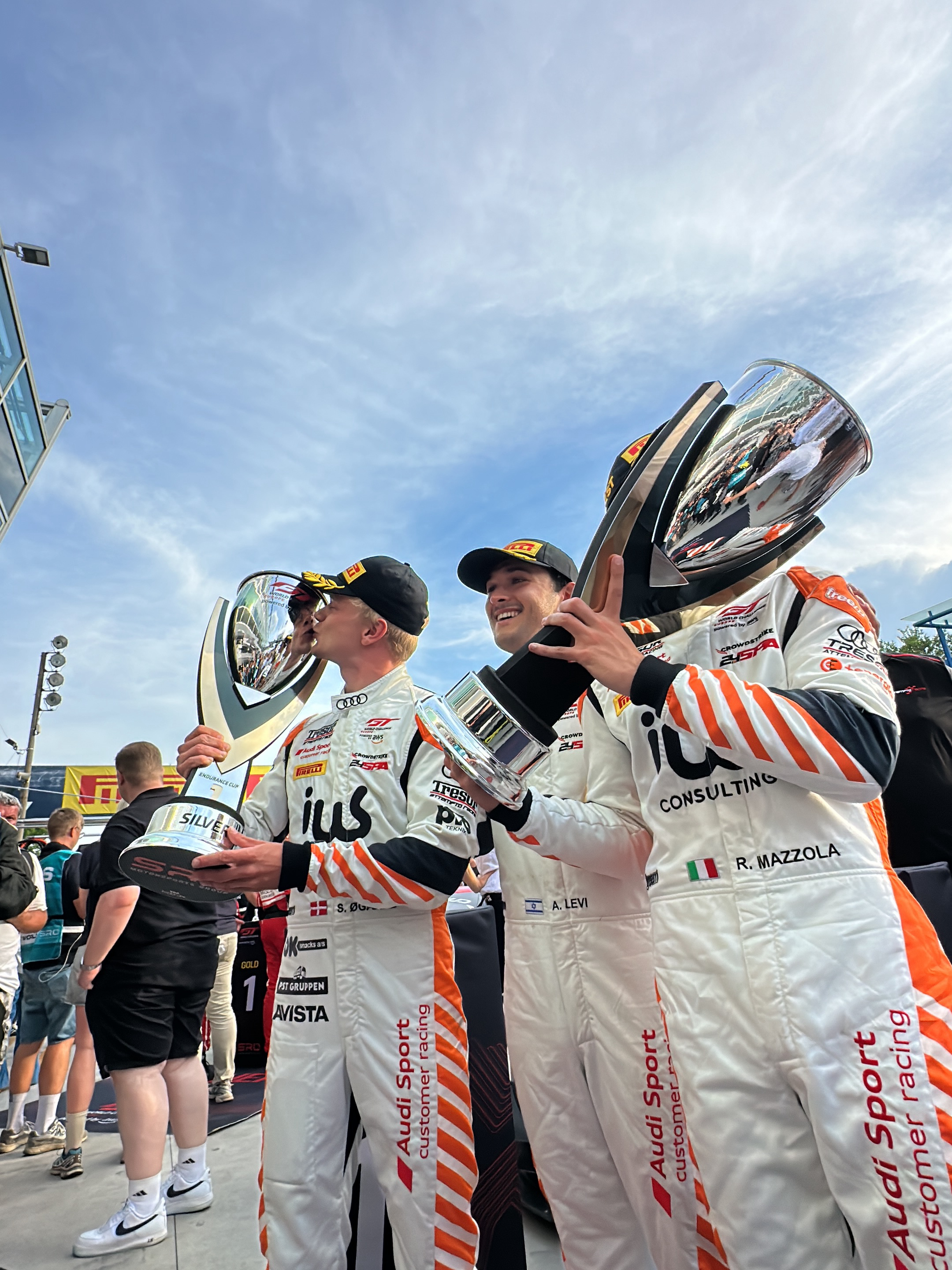 Three race car drivers in white uniforms with orange stripes stand on a podium holding trophies. The driver on the left is kissing his trophy, marked "Silver." The driver in the middle holds a trophy marked with an Israeli flag, and the driver on the right holds a larger trophy. The uniforms display "Audi Sport Customer Racing" logos and country flags. The name "R. Mazzola" is visible on the right driver's uniform with an Italian flag. The environment includes a building with glass panels and a cloudy sky, consistent with the city of Monza, Italy, where the Autodromo Nazionale Monza is located. Other individuals are seen in the background.