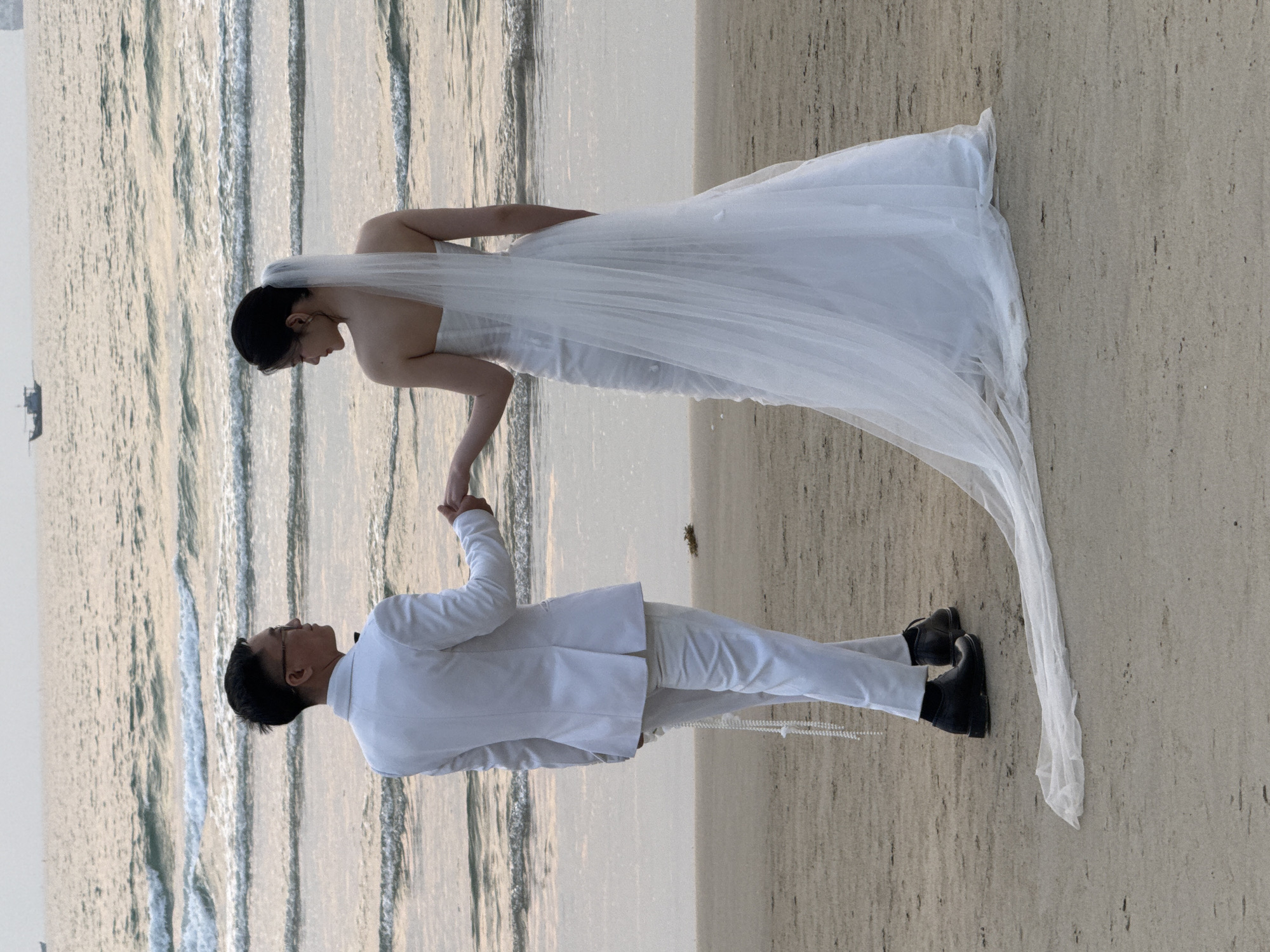 A wedding couple is standing on a beach in Đà Nẵng, Vietnam. The bride is wearing a white wedding dress and veil, while the groom is wearing a white suit and bow tie. They are holding hands and looking at each other. The beach is sandy, and the water is calm. The image has a soft, romantic feel.
