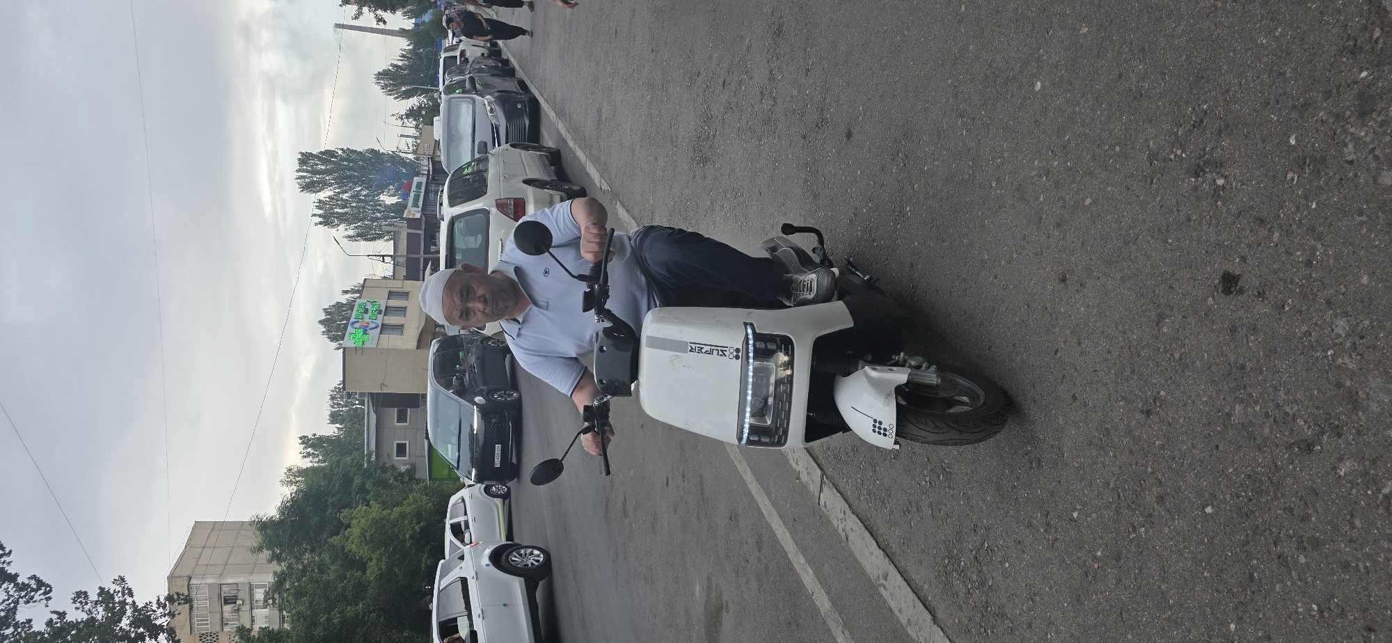 A middle-aged man wearing a light blue polo shirt, dark jeans, and a white skullcap rides a white electric scooter down an asphalt road in Bishkek, Kyrgyzstan. He is looking directly at the camera with a neutral expression. The scooter has a white body with some grey stripes and a rectangular LED headlight. The background shows a street with parked cars, including a white SUV and several vans, and buildings with residential windows and commercial signs, one of which appears to be a pharmacy with a green cross logo. The sky is overcast.