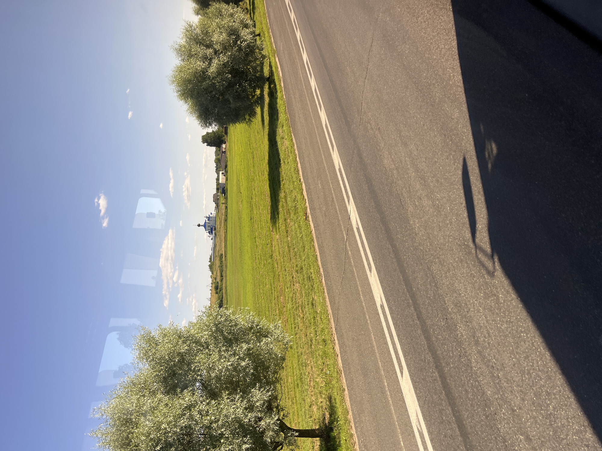 A rural landscape is depicted under a bright blue sky with scattered white clouds. A paved road with double white lines runs diagonally across the foreground, casting a long shadow to the right. To the left of the road, a verdant green field stretches into the distance, bordered by a line of trees and a small cluster of buildings. In the middle ground, a white church with a blue dome and cross stands prominently, suggesting a location near Myadzyel, Belarus. Two large, leafy trees frame the scene on either side, their foliage a light green. The overall impression is one of a peaceful, sunny day in the countryside.