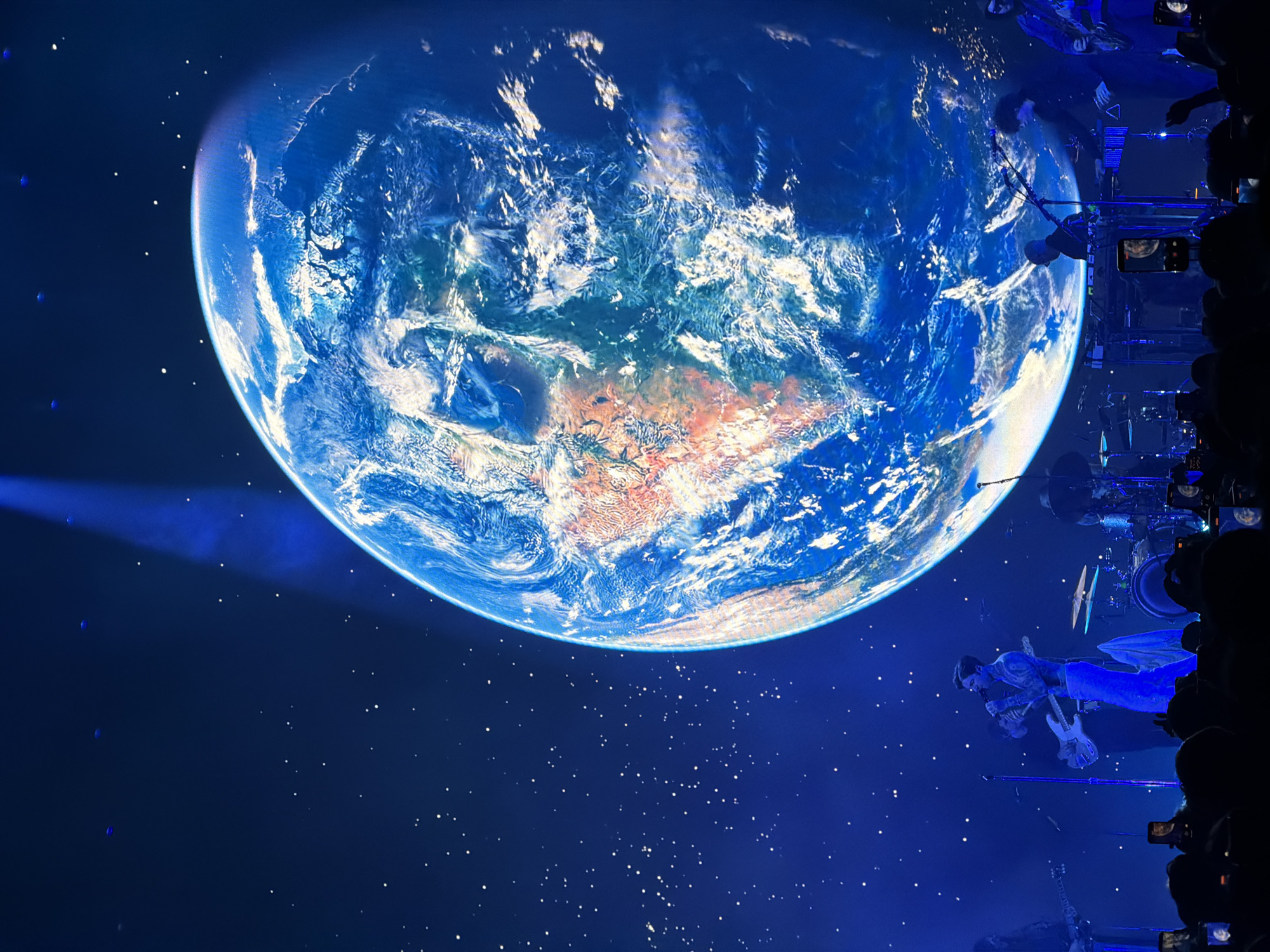 The image depicts a concert setting, potentially at the O2 Arena in London, as evidenced by the high production value and large-scale staging. A massive screen displays a view of Earth, centered on Australia, against a backdrop of simulated stars. Performers are suspended from rigs in the air, with musical instruments visible. Spectators hold up phones to capture the event, with their silhouettes visible along the edges of the image. The lighting is primarily blue, emphasizing the celestial theme.