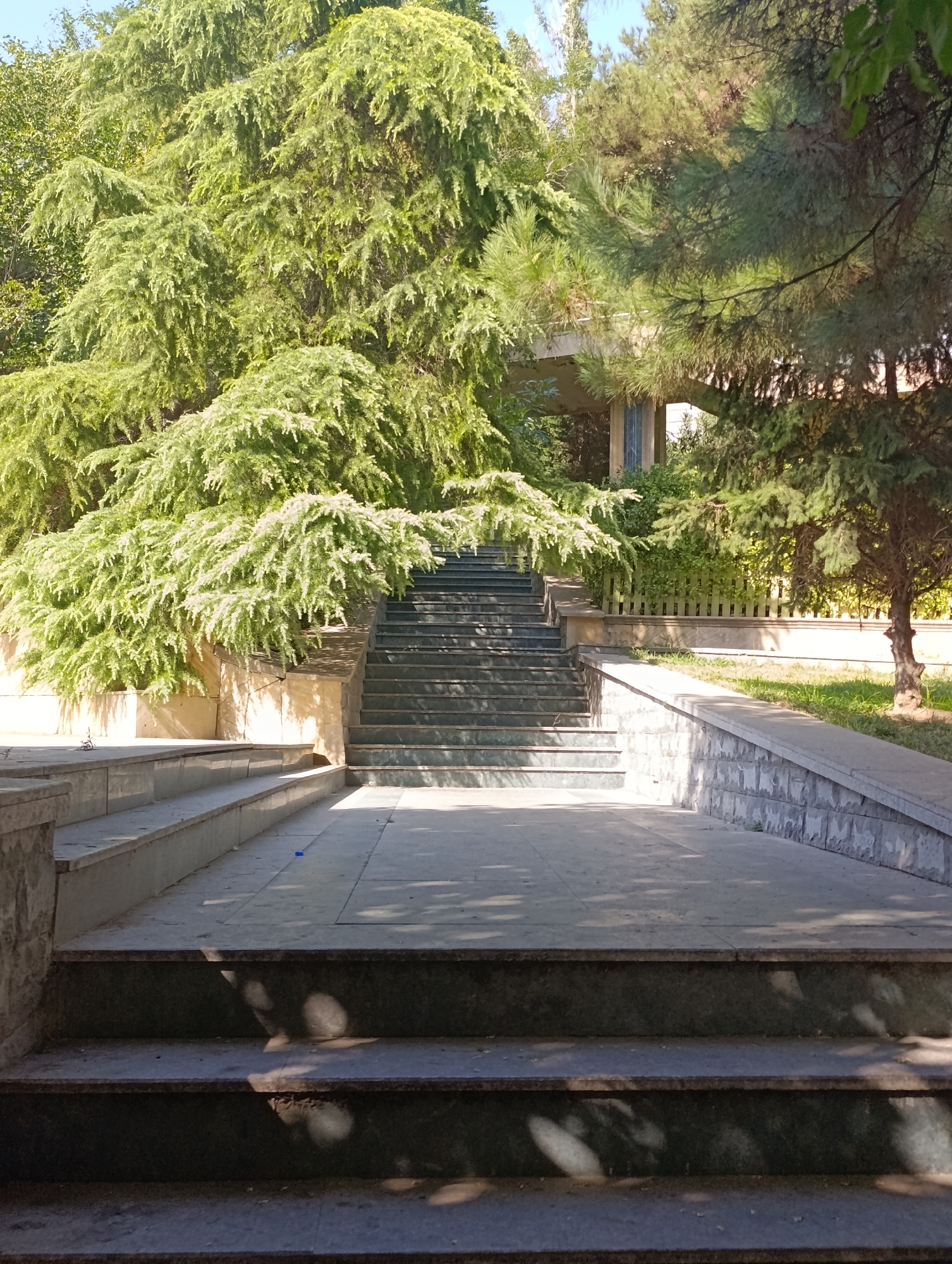 The image depicts an outdoor scene with a set of stone stairs leading upwards, flanked by lush green trees. In the background, a bridge-like structure is partially visible above the trees. The stairs are made of dark stone and are well-maintained, with some shadows cast upon them by the surrounding foliage. The overall impression is one of a serene, possibly park-like setting. The presence of trees and the architectural style suggest a location that could be found in a city with a mix of natural and built environments. Given the mention of Baku, Azerbaijan, this scene might be found in one of its many parks or public spaces, possibly near an elevated walkway or overpass.