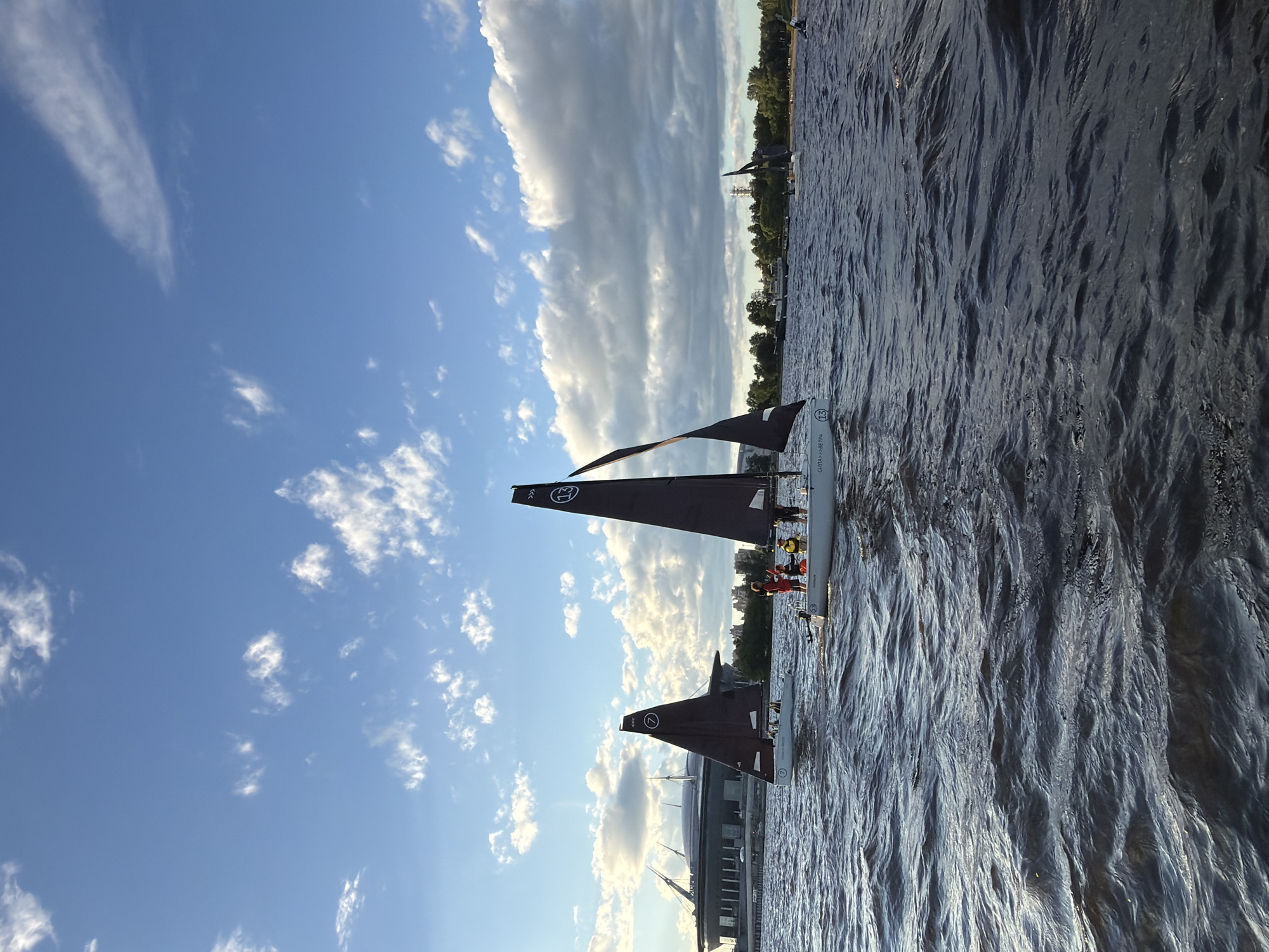 In Saint Petersburg, Russia, on a body of water, several small sailboats with dark sails are visible. A modern stadium with a domed roof is situated in the background, to the left of the frame. The sky is a clear blue with scattered white clouds. The water is choppy, indicating wind. On the closest sailboat, there are at least three individuals visible, appearing to be engaged in sailing activities. Another sailboat to the left also has individuals on board. The overall scene suggests a recreational sailing event or lesson.