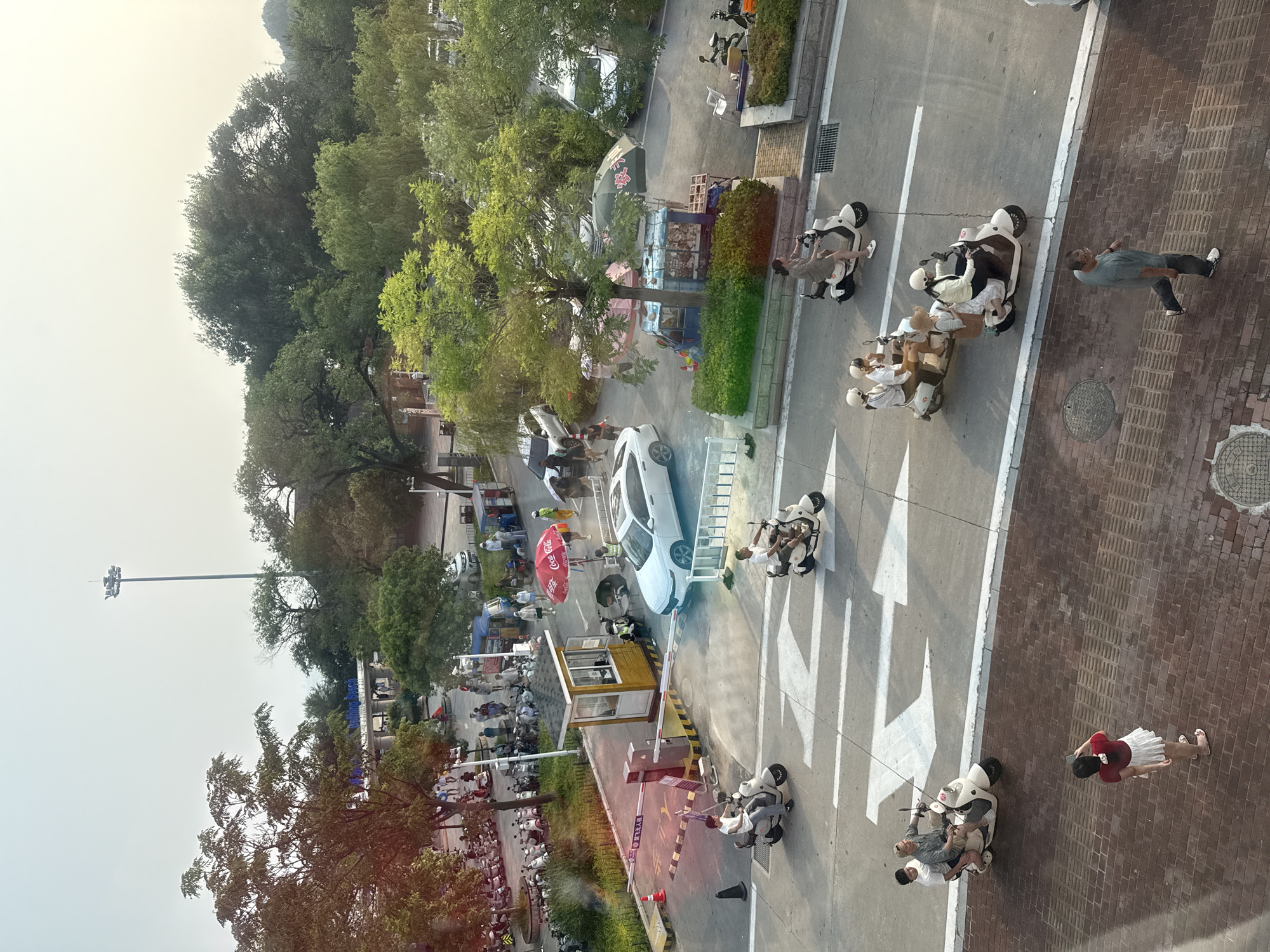 The image depicts a street scene near a beach in Qinhuangdao, China. In the foreground, a brick-paved walkway is visible, with a man walking from right to left. Further up, a woman in a red shirt and white skirt is standing and taking a photo with her phone. The street in the middle ground is paved with asphalt and marked with white directional arrows. Several electric scooters are navigating the street. One scooter carries two men, another carries three people, and two more scooters carry two people each, with passengers wearing helmets. On the right side of the street, a parked scooter and two people are near a stall with a green umbrella.
In the background, a white sedan is parked near a small booth with a yellow roof, likely a toll or entry booth for a parking area. A red Coca-Cola umbrella shades a seating area. Beyond this, a row of parked scooters is visible, suggesting a public transportation hub or rental area. Trees line the background, and a tall light pole stands prominently. In the distance, a sign with blue lettering reads "Bathing Beach." The sky is overcast with a yellowish tint, indicating either dawn, dusk, or hazy conditions. The overall scene suggests a busy public area with a mix of transportation methods and commercial activity.