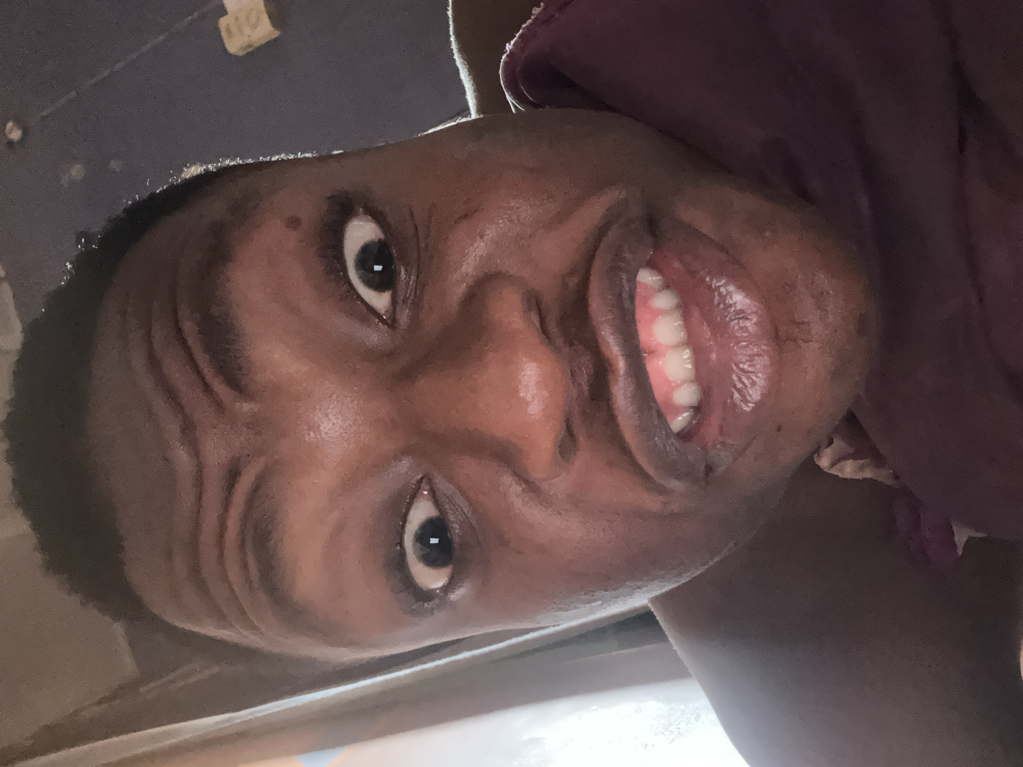 A close-up, rotated image shows a person with dark skin. Their mouth is open, displaying teeth. The individual is wearing a dark purple garment. The background is indistinct, with a portion appearing to be part of a vehicle. The location is Unknown, Unknown.